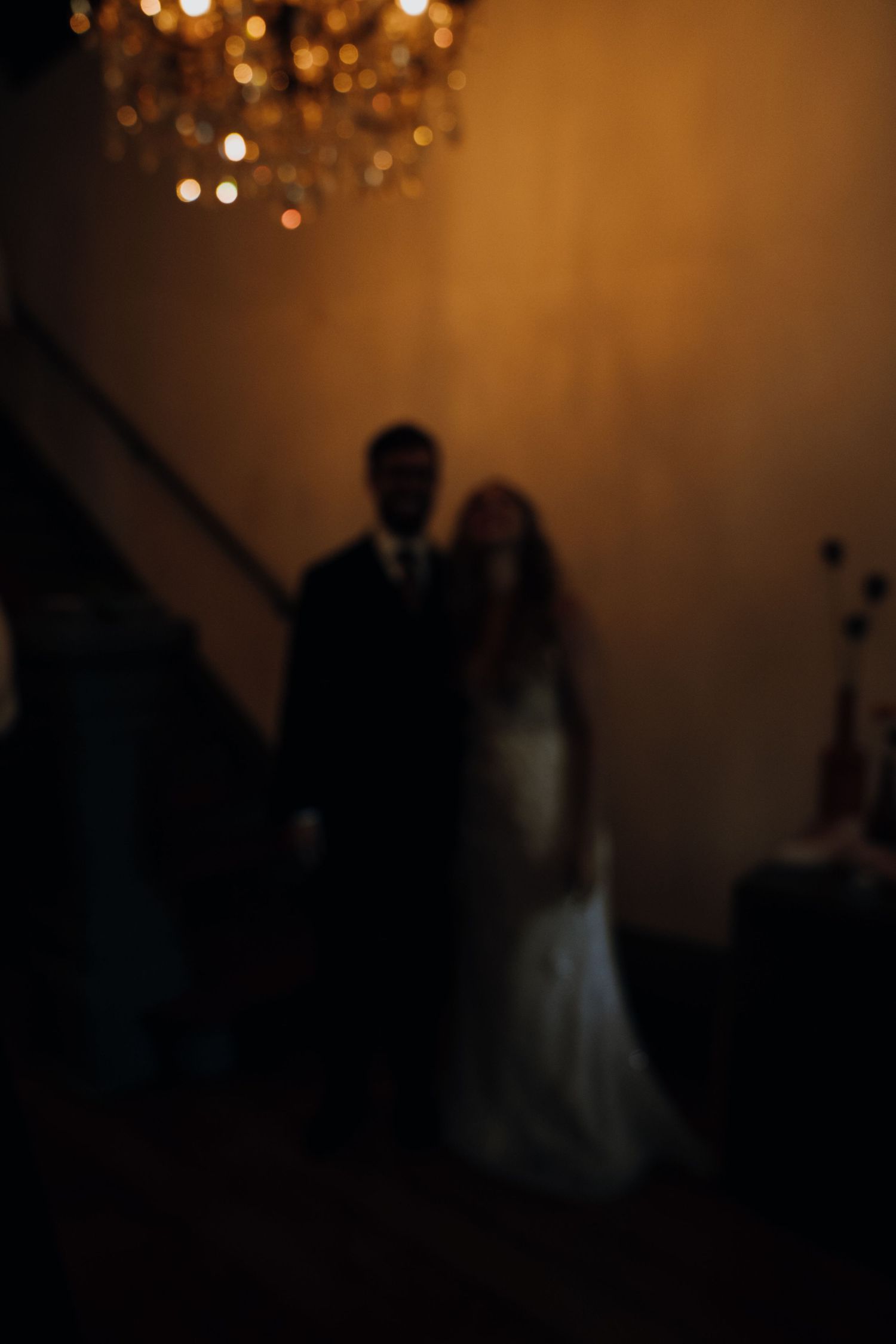 Bride and groom silhouetted under a glowing chandelier in a dimly lit wedding venue.