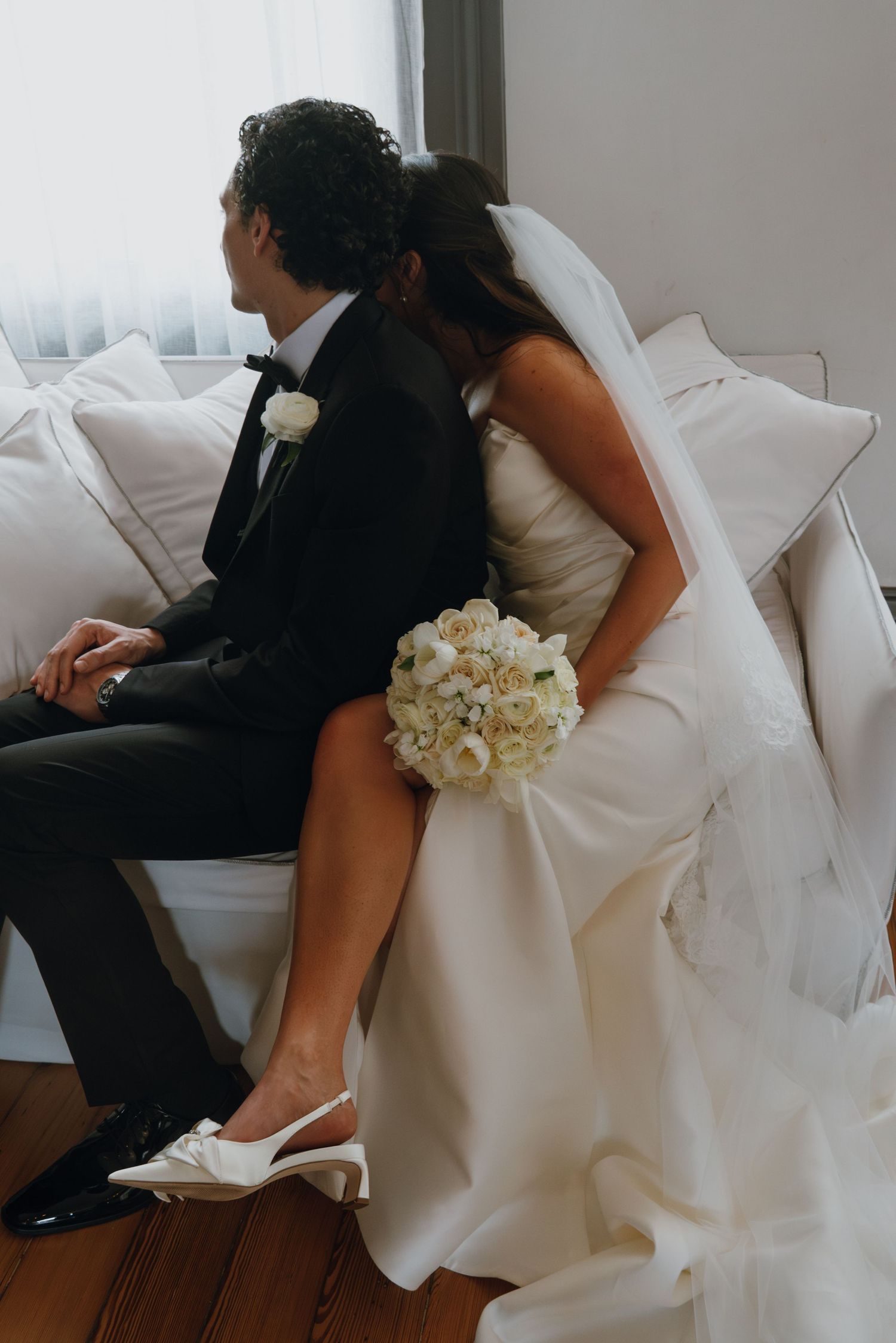 Bride and groom sitting back-to-back on white sofa, bride holding white rose bouquet wearing slingback heels.