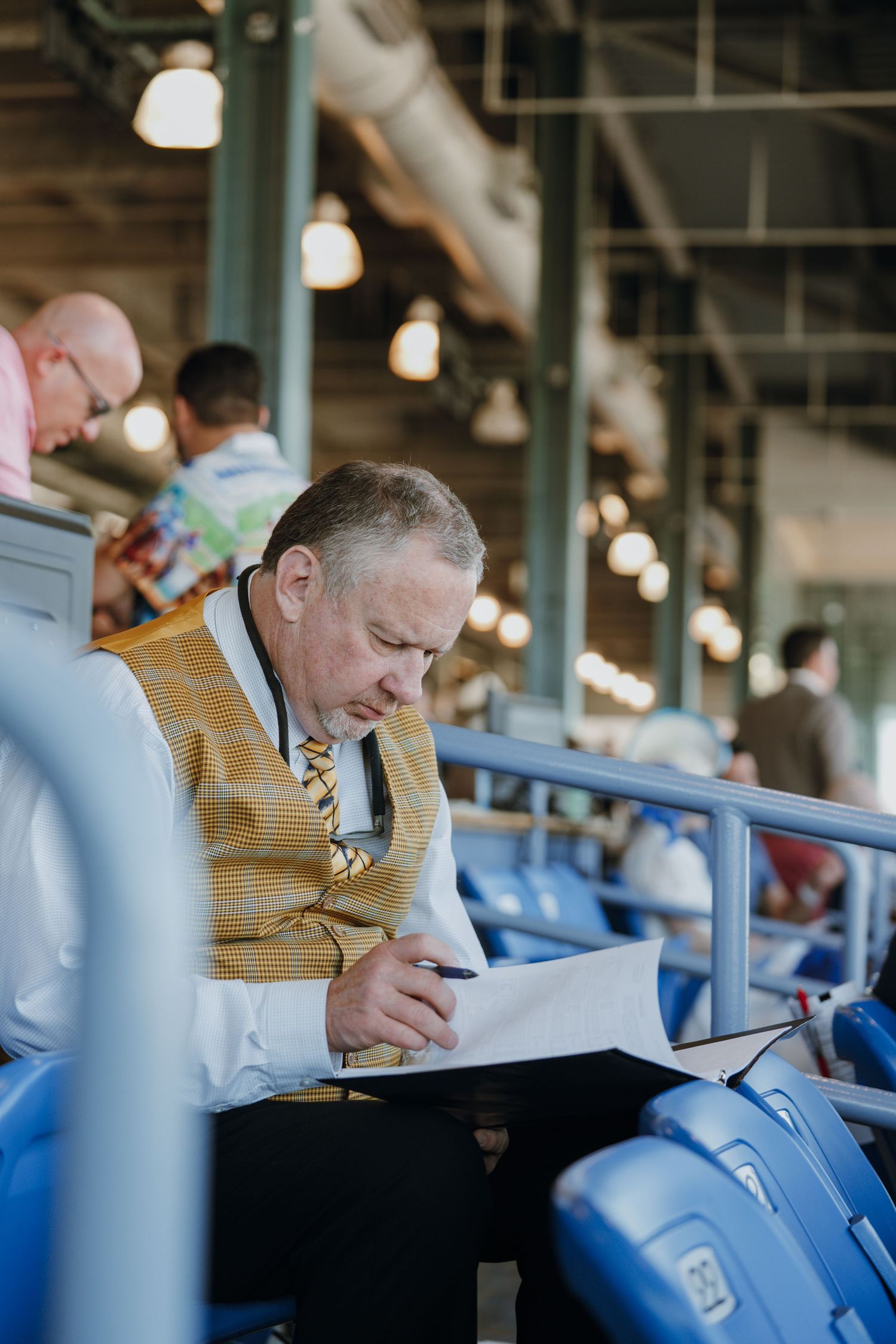 Man in striped vest writes notes in stadium seating at a baseball game.