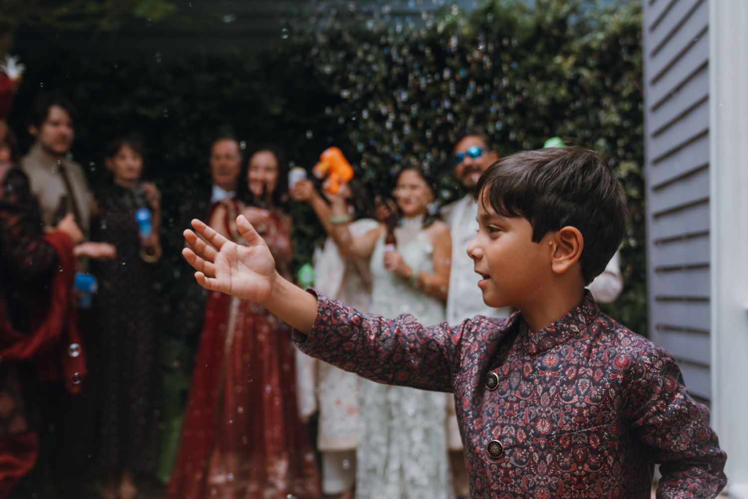 Young boy in traditional Indian attire reaching out to catch bubbles at a wedding celebration.