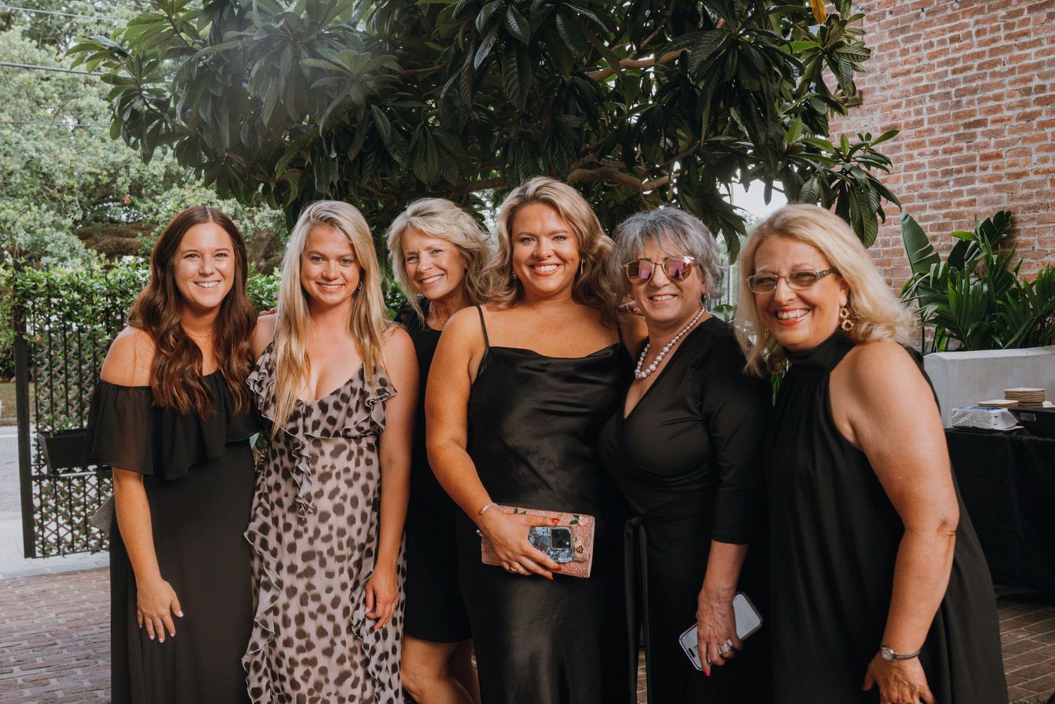 Six women in black dresses pose together smiling at an outdoor evening event.