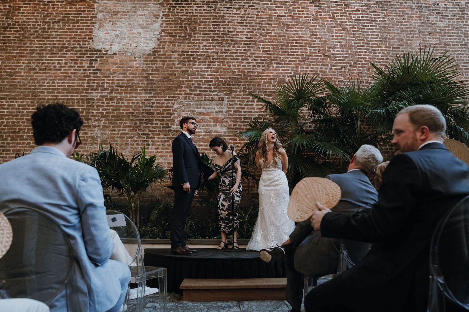 Bride and groom exchange vows before an officiant against a rustic brick wall with tropical plants.