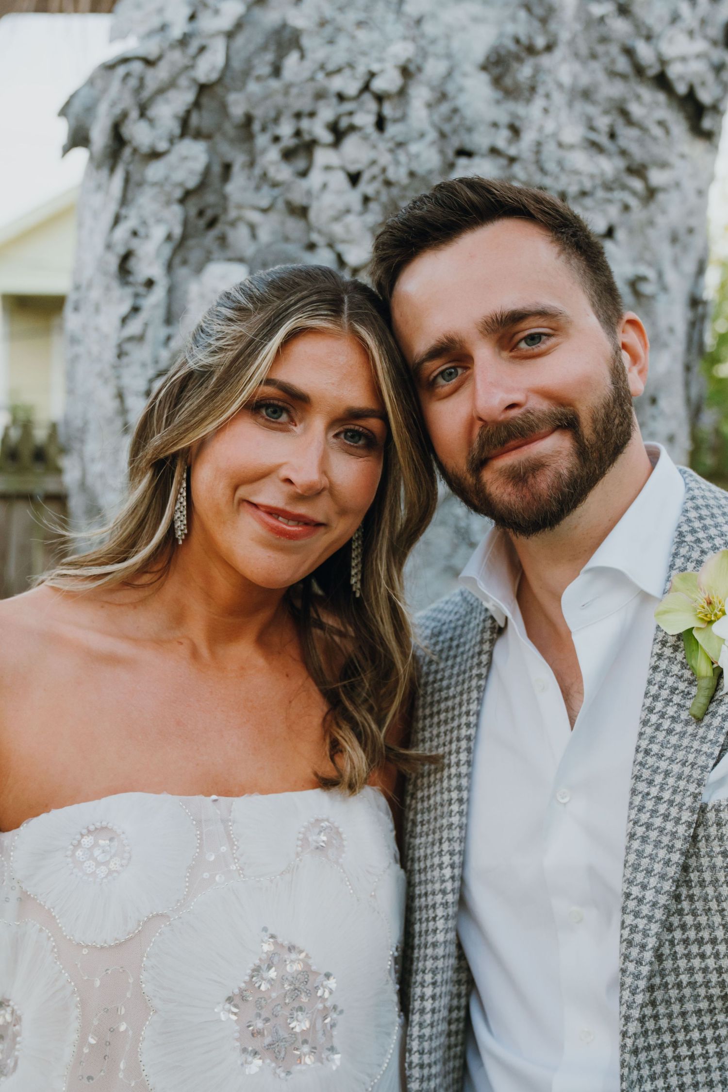 Portrait of smiling bride with silver drop earrings and groom in grey houndstooth blazer with white boutonniere.