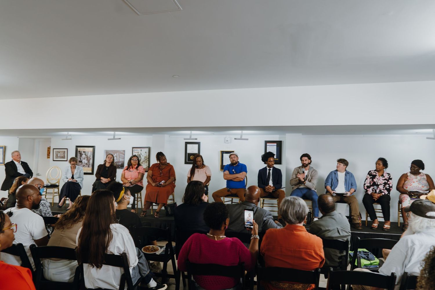 A large community panel discussion with multiple speakers seated facing an engaged audience in a bright room.