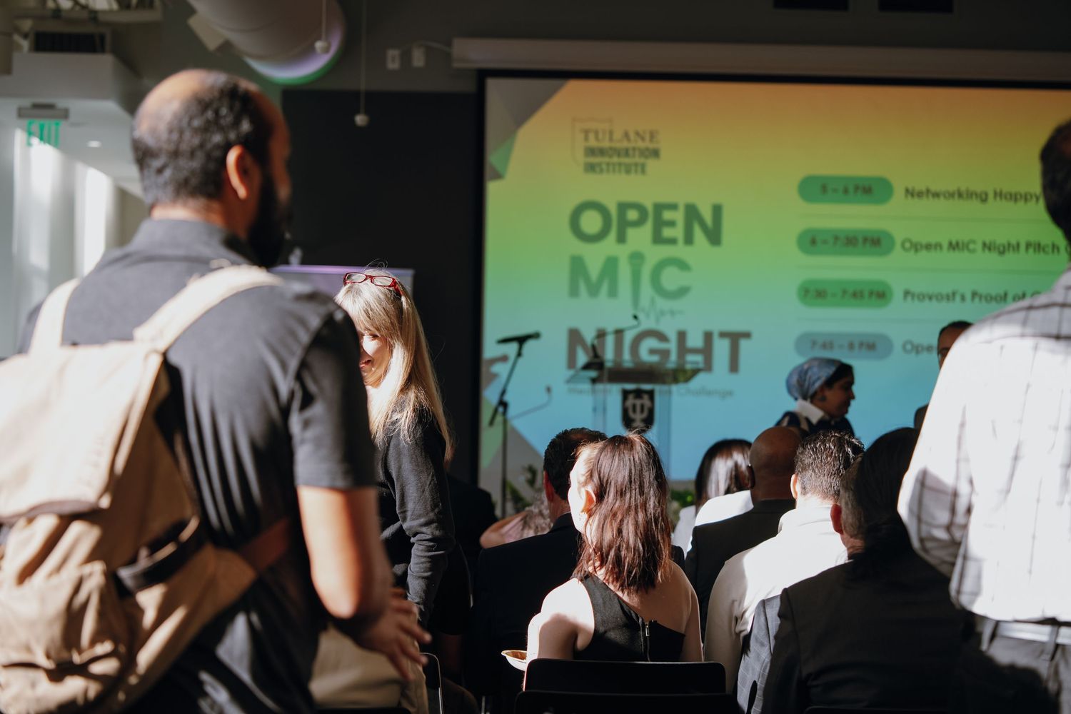 Audience watches Open Mic Night presentation at Tulane Innovation Institute event with colorful screen display.
