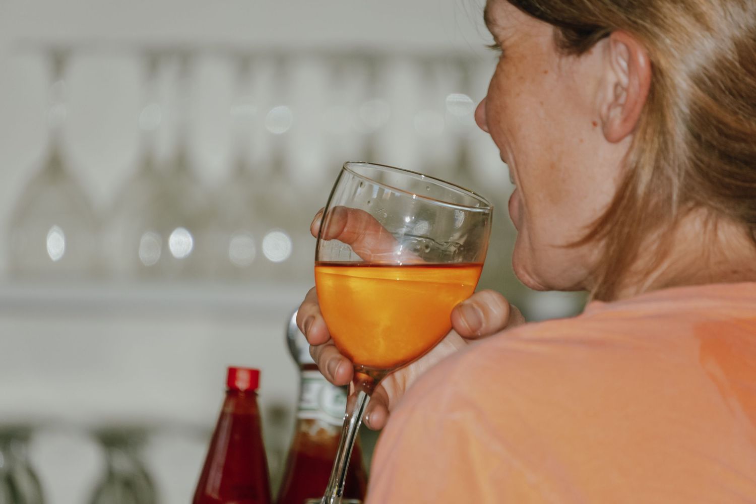 Woman in peach shirt drinking orange cocktail in wine glass at a bar with bottle display.