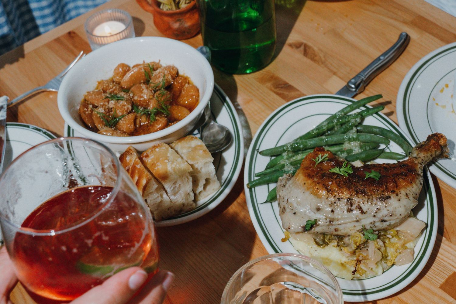 Restaurant table with roast chicken, green beans, potatoes, and bread with a cocktail in foreground.