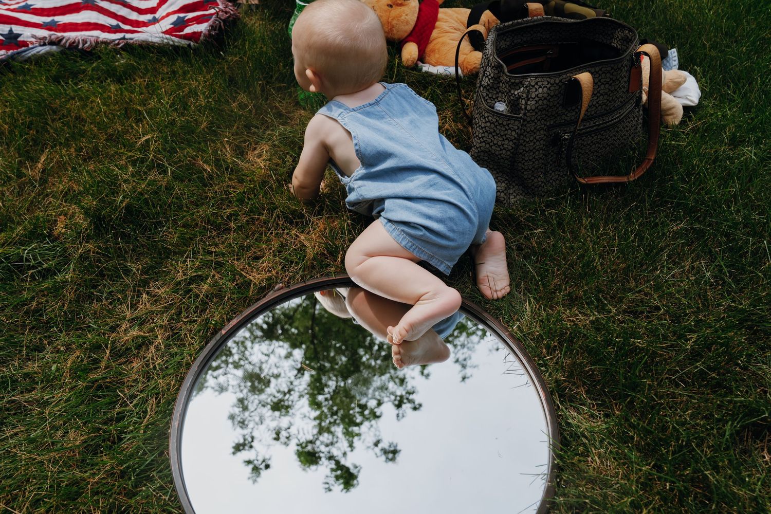 A baby in a blue romper crawls toward a round mirror on green grass beside a picnic basket.