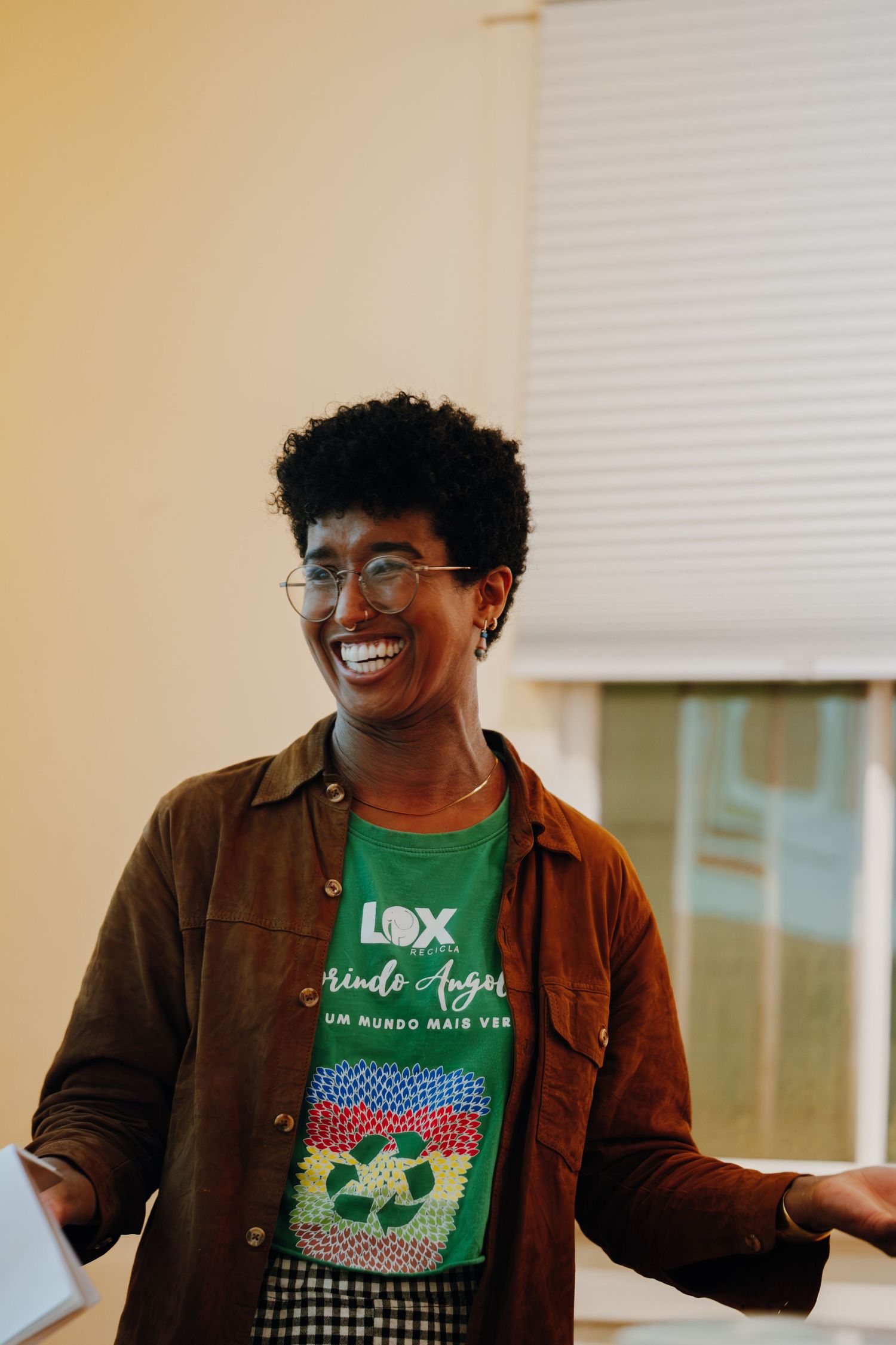 Smiling person wearing glasses and a green LOX t-shirt with a brown jacket, laughing joyfully indoors.
