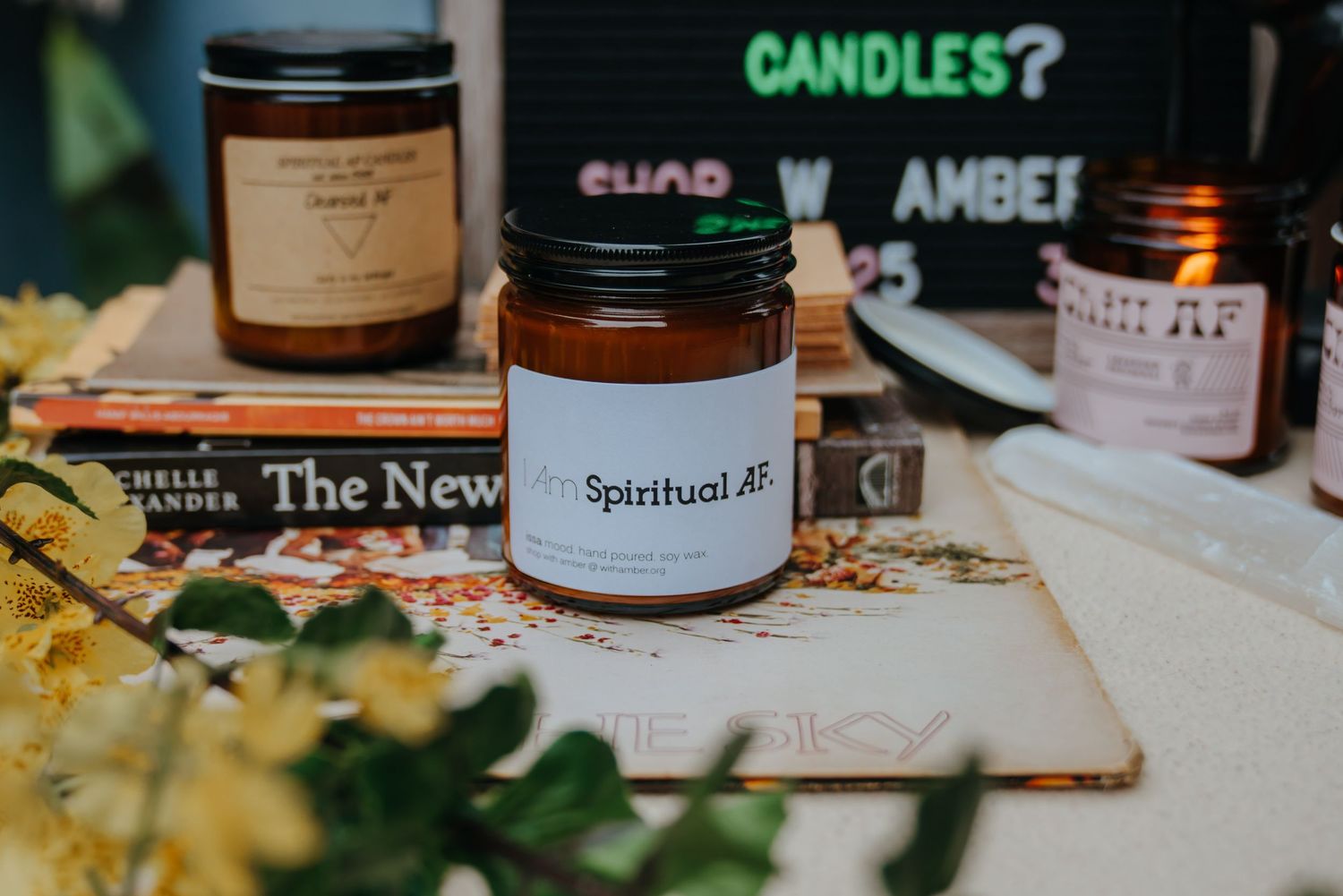 Amber glass Spiritual AF scented candle jar displayed on books surrounded by yellow flowers and candles in a cozy setting.