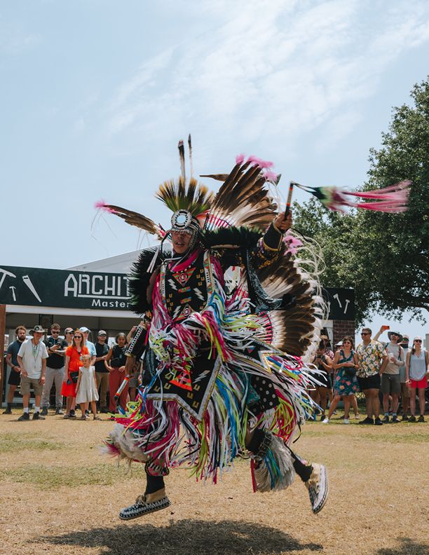 Native American dancer in vibrant traditional regalia performs at an outdoor powwow ceremony before a crowd of spectators.
