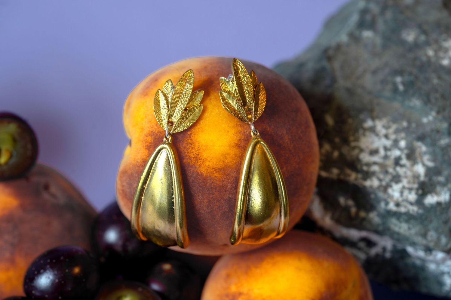 Gold leaf and teardrop dangle earrings displayed on a fresh peach with dark fruits.