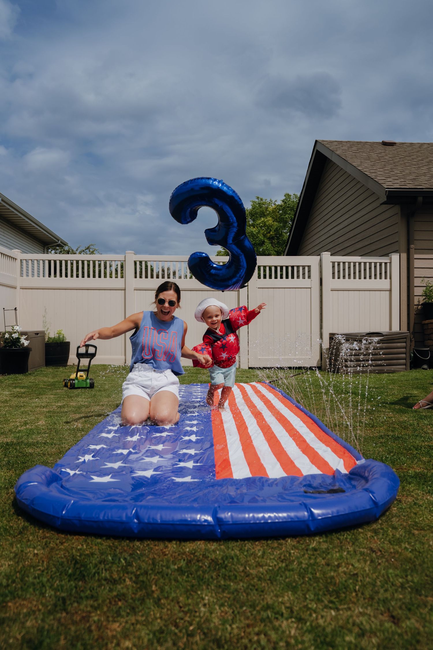 Mother and child celebrating a 3rd birthday on an American flag slip-and-slide in backyard.