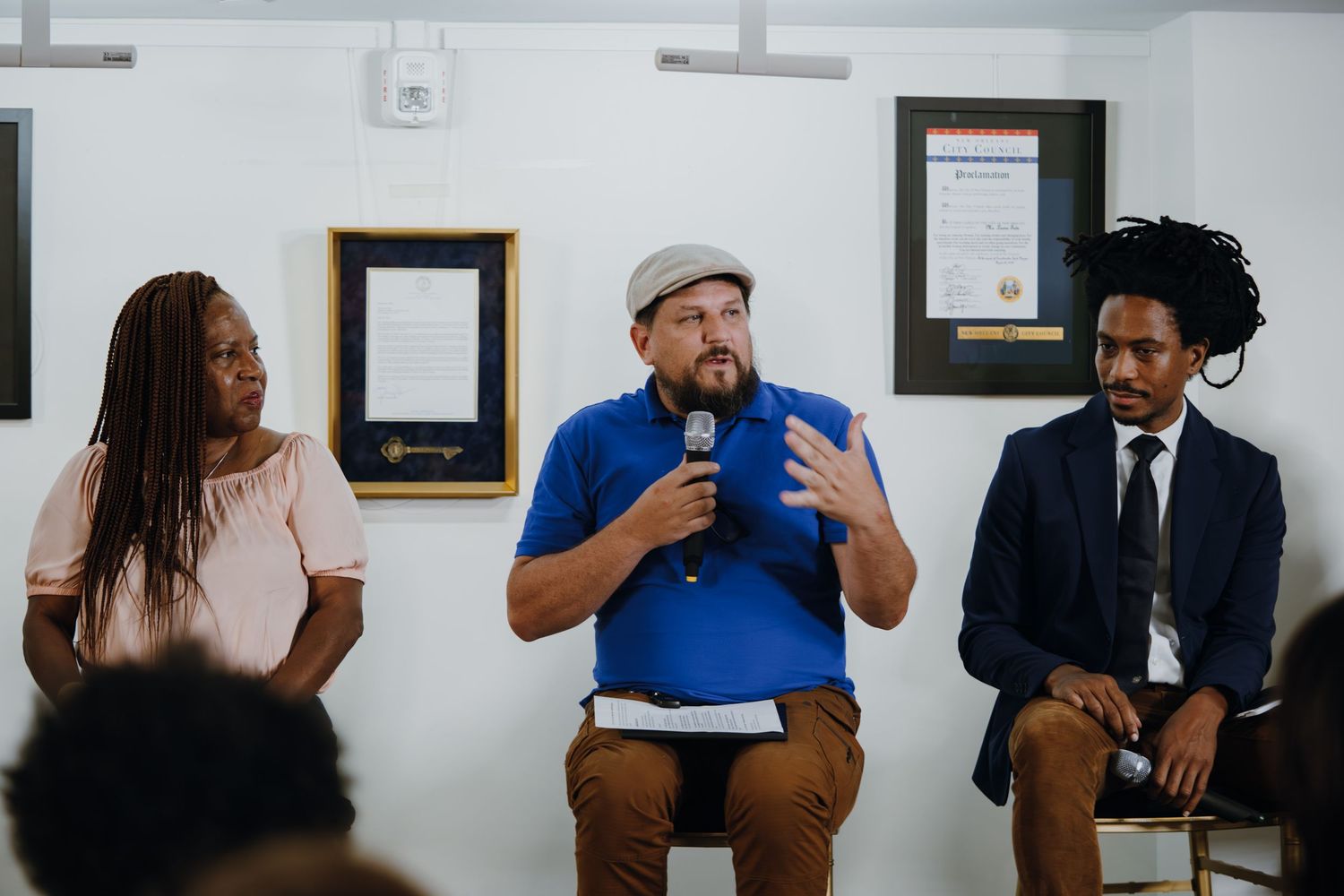 Three panelists seated at a community forum, with a man in a blue shirt and cap speaking into a microphone.