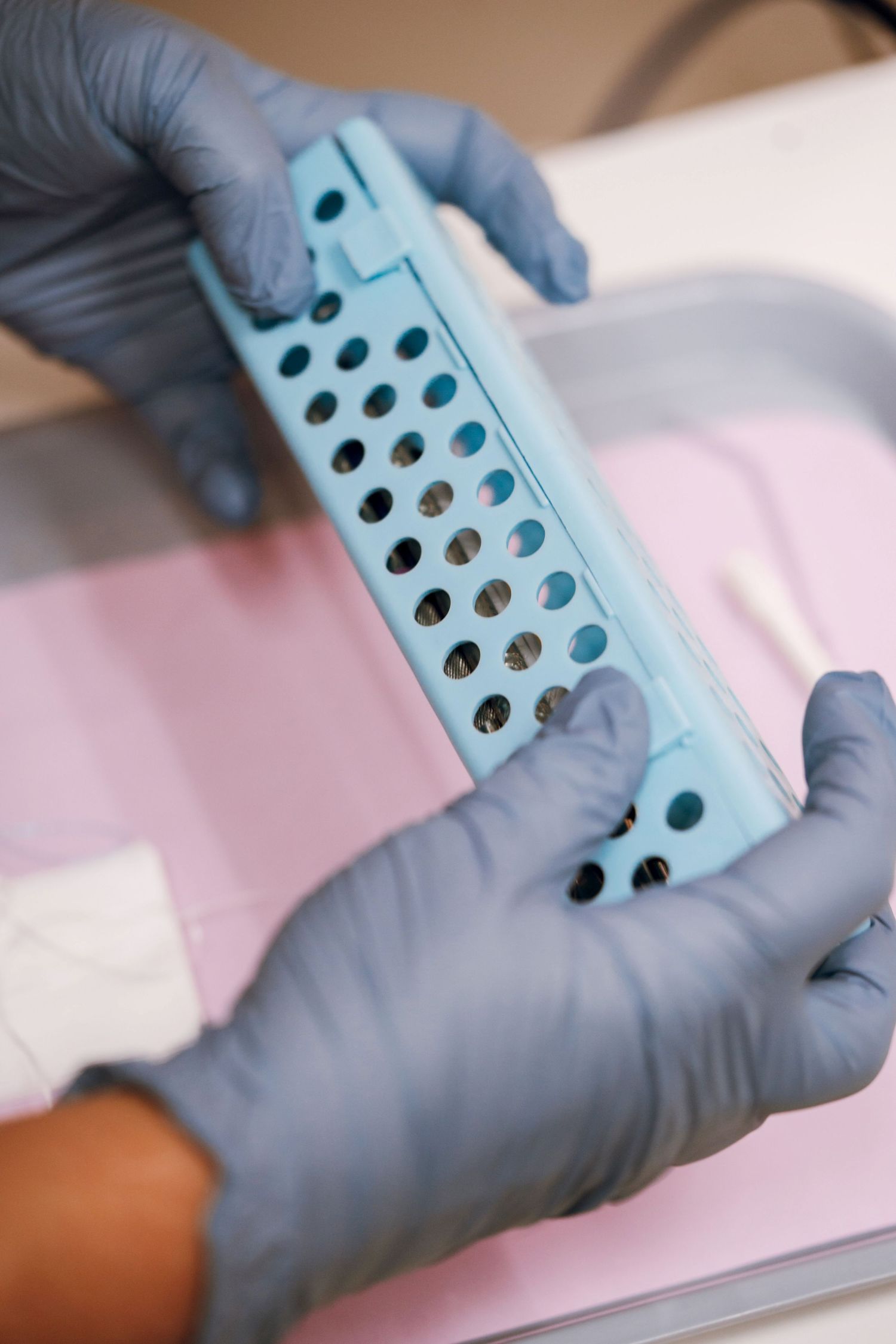 Gloved hands hold a blue sterilization tray with holes over a pink dental tray.