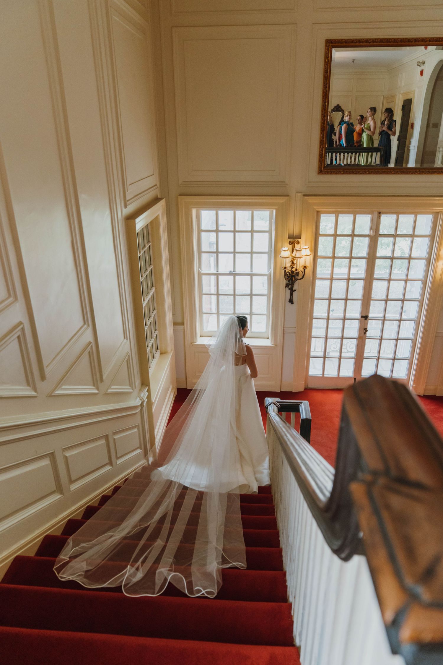 Bride in a flowing white gown with long veil descending red-carpeted staircase in elegant venue. New Orleans, Louisiana, Mississippi