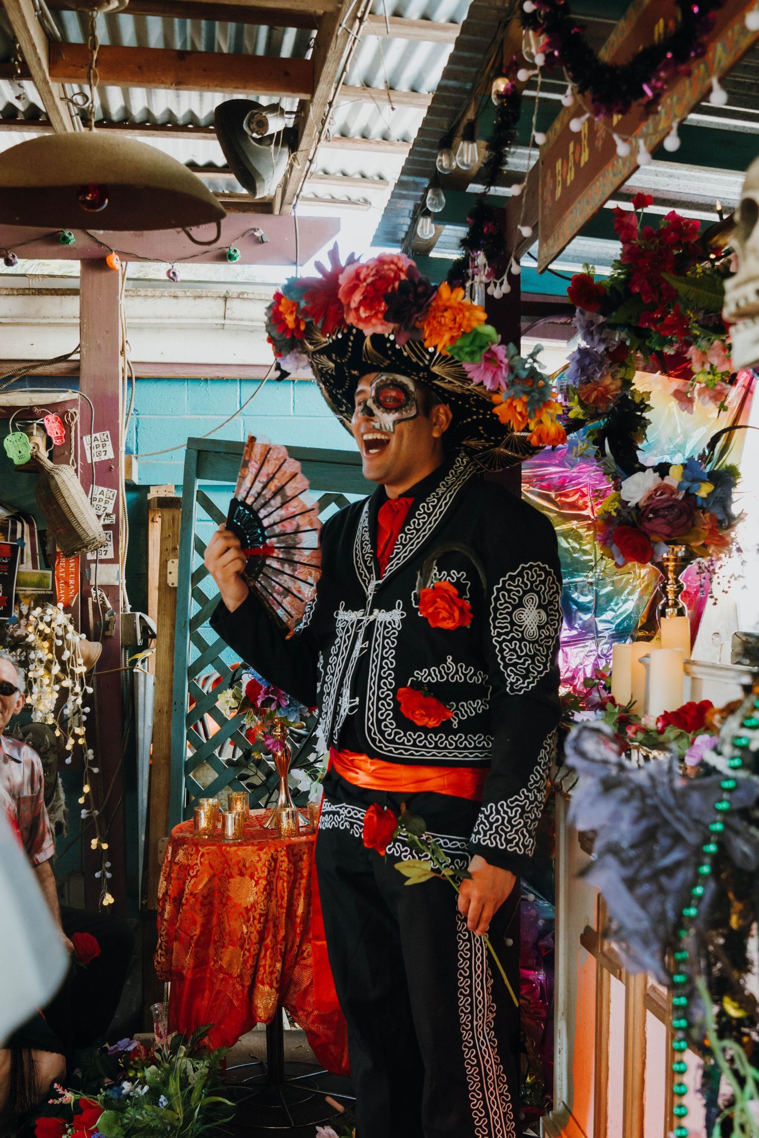 Man in Dia de los Muertos mariachi costume with sugar skull makeup holds a fan at a colorful festival.