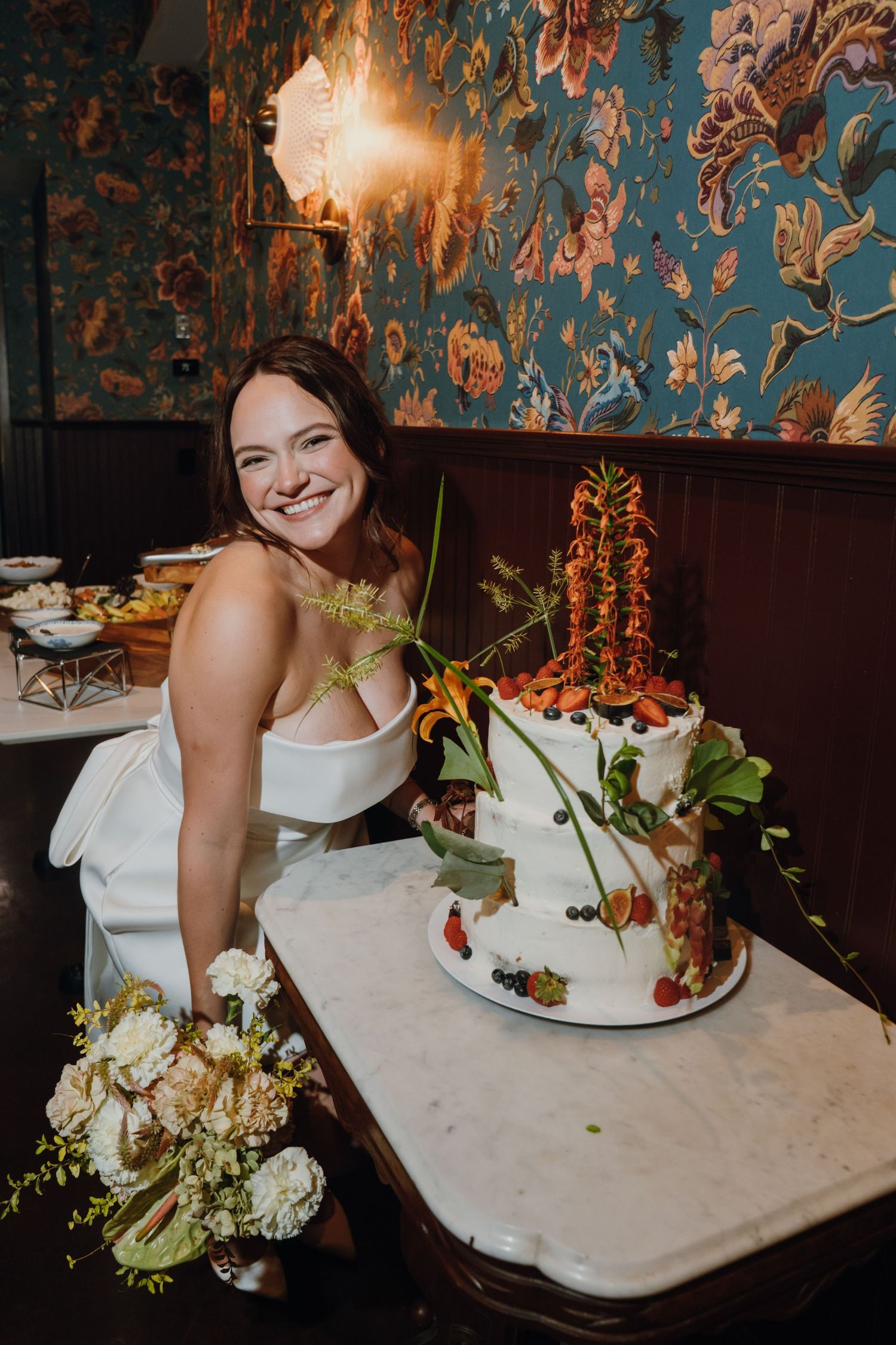 Smiling bride in white strapless gown poses with floral bouquet beside decorated two-tier wedding cake with fresh fruit. New Orleans, Louisi