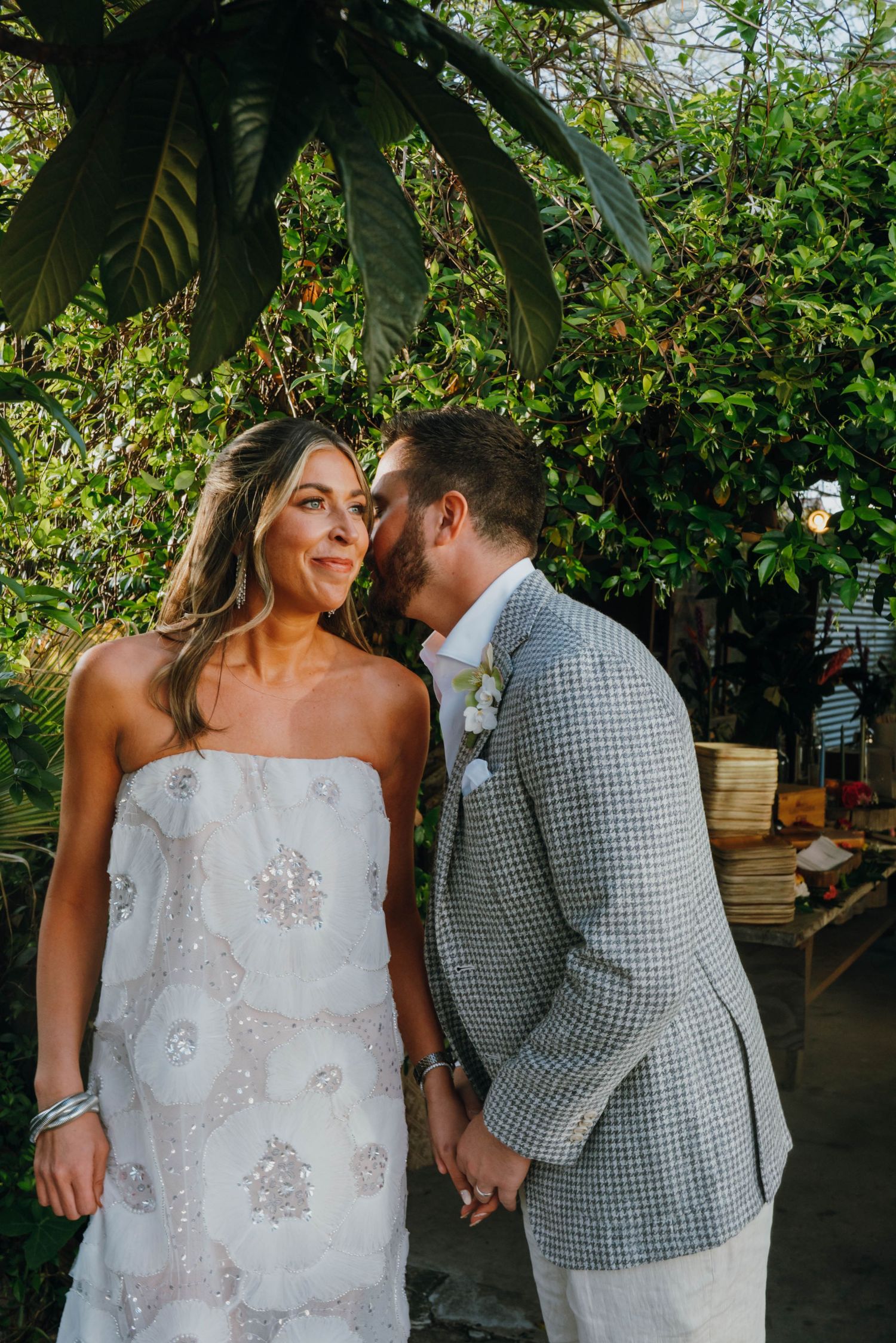 Groom in grey houndstooth blazer whispers to smiling bride in strapless white beaded gown at garden wedding.