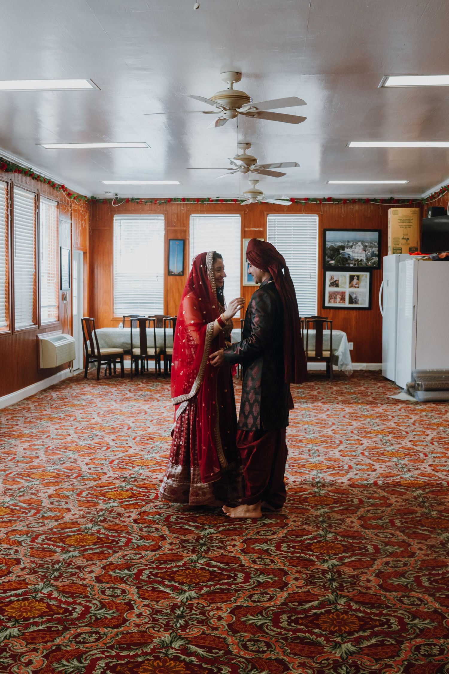 Indian bride and groom in traditional red and dark attire sharing an intimate moment on ornate carpet.
