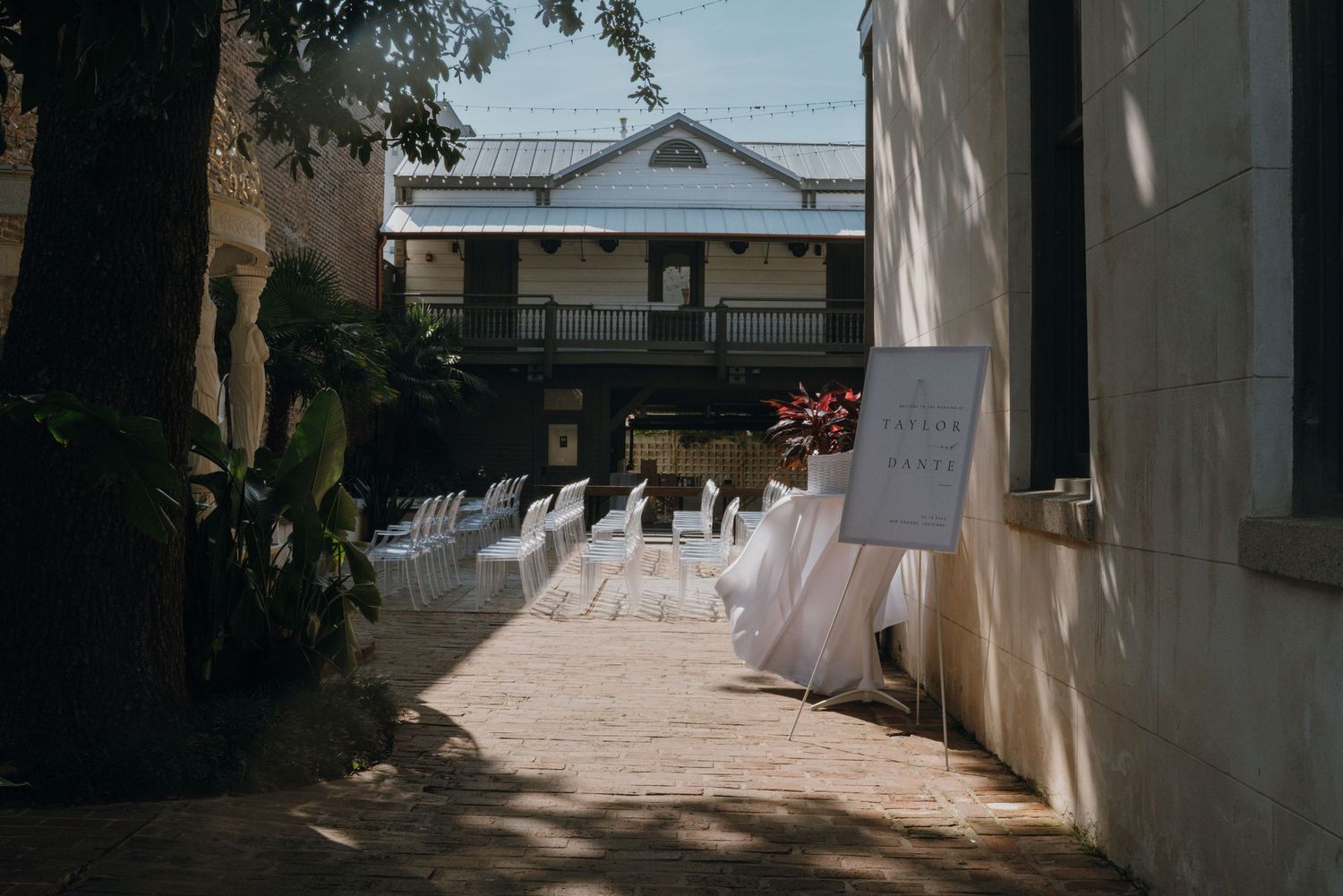 Sunlit outdoor wedding ceremony aisle with white chairs, welcome sign, and historic building in background.