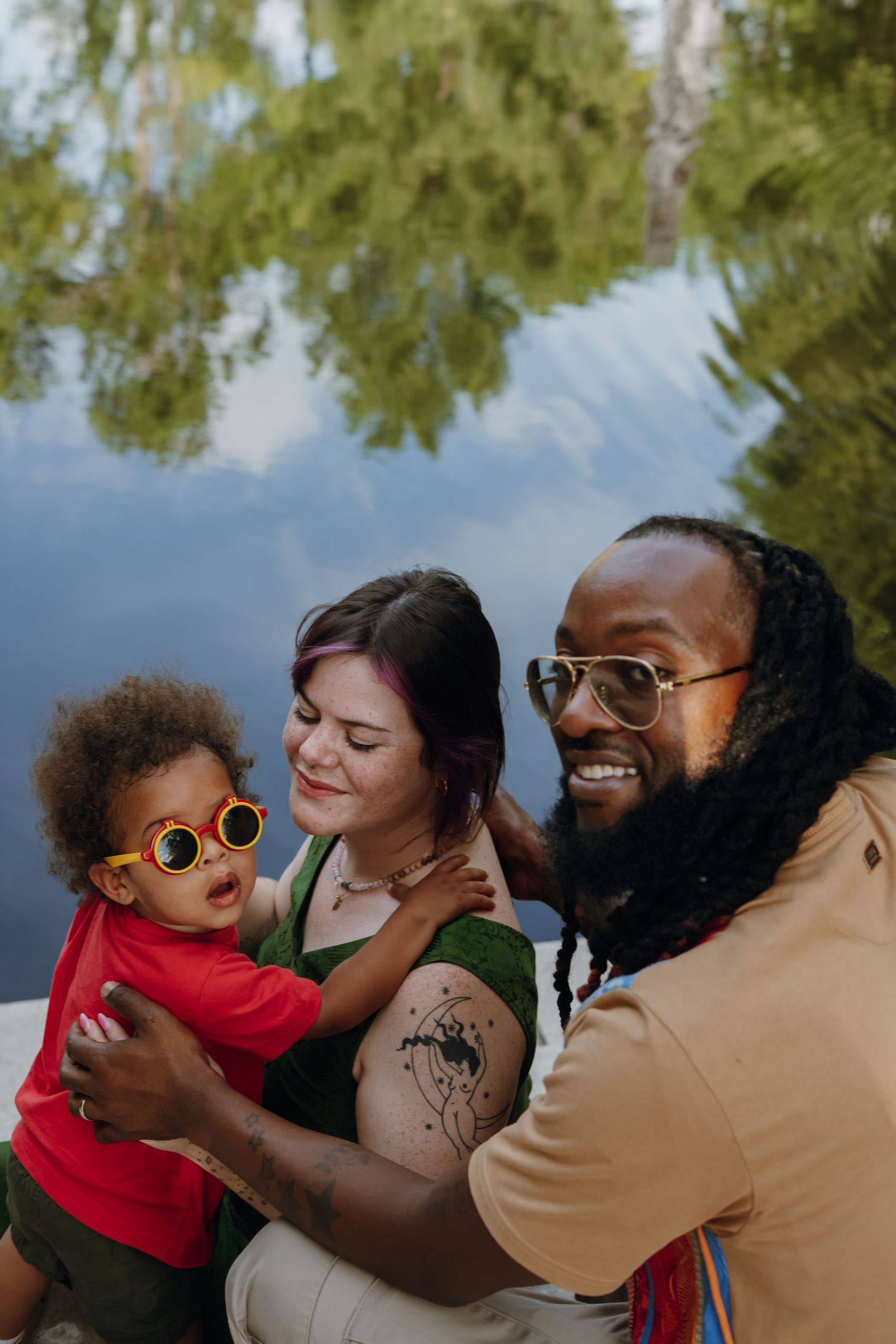 A joyful multiracial family with a baby wearing yellow sunglasses poses outdoors near a reflective pond.