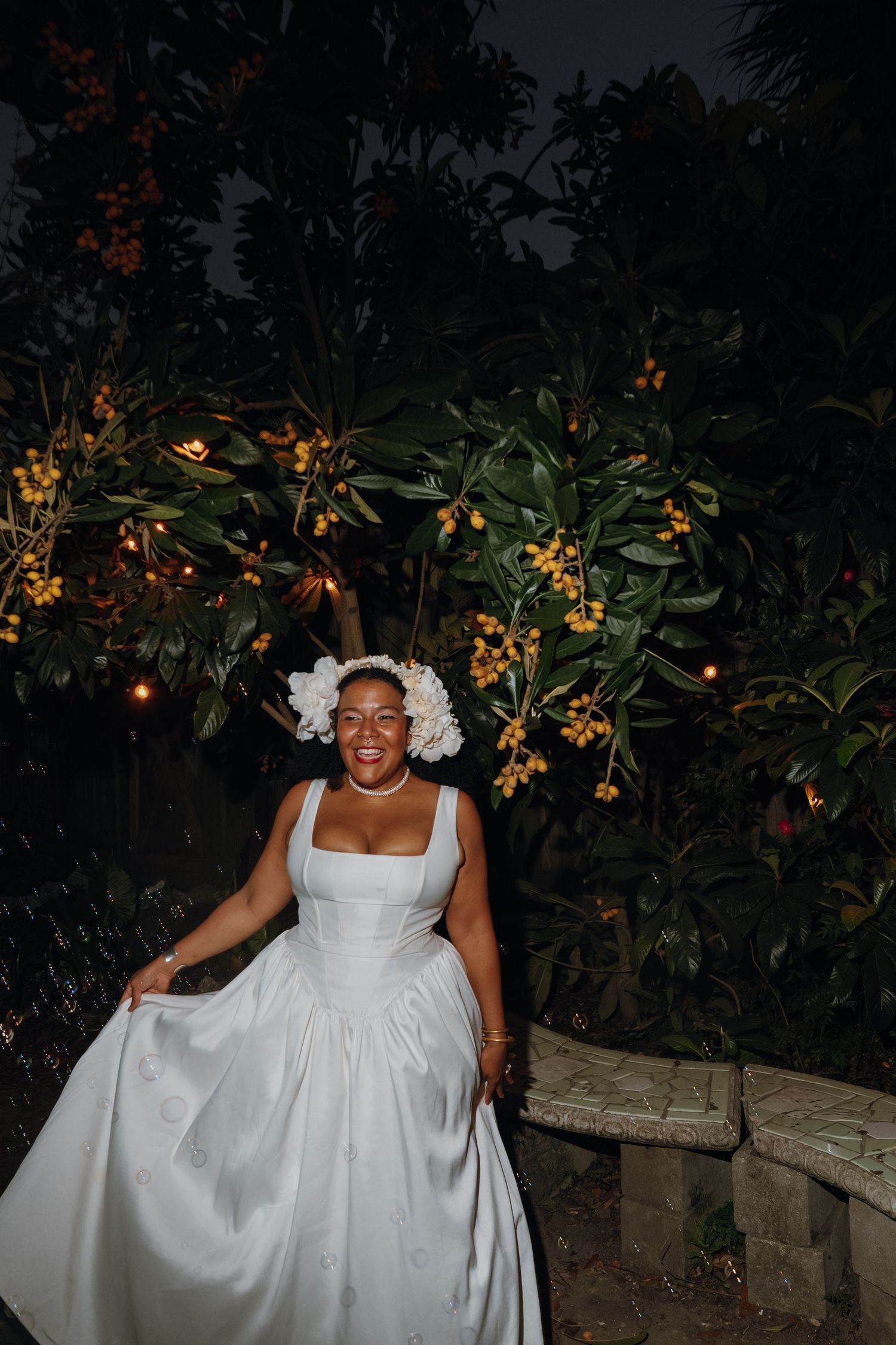 Joyful bride in white ball gown holding skirt poses before fruit tree with warm string lights at dusk.