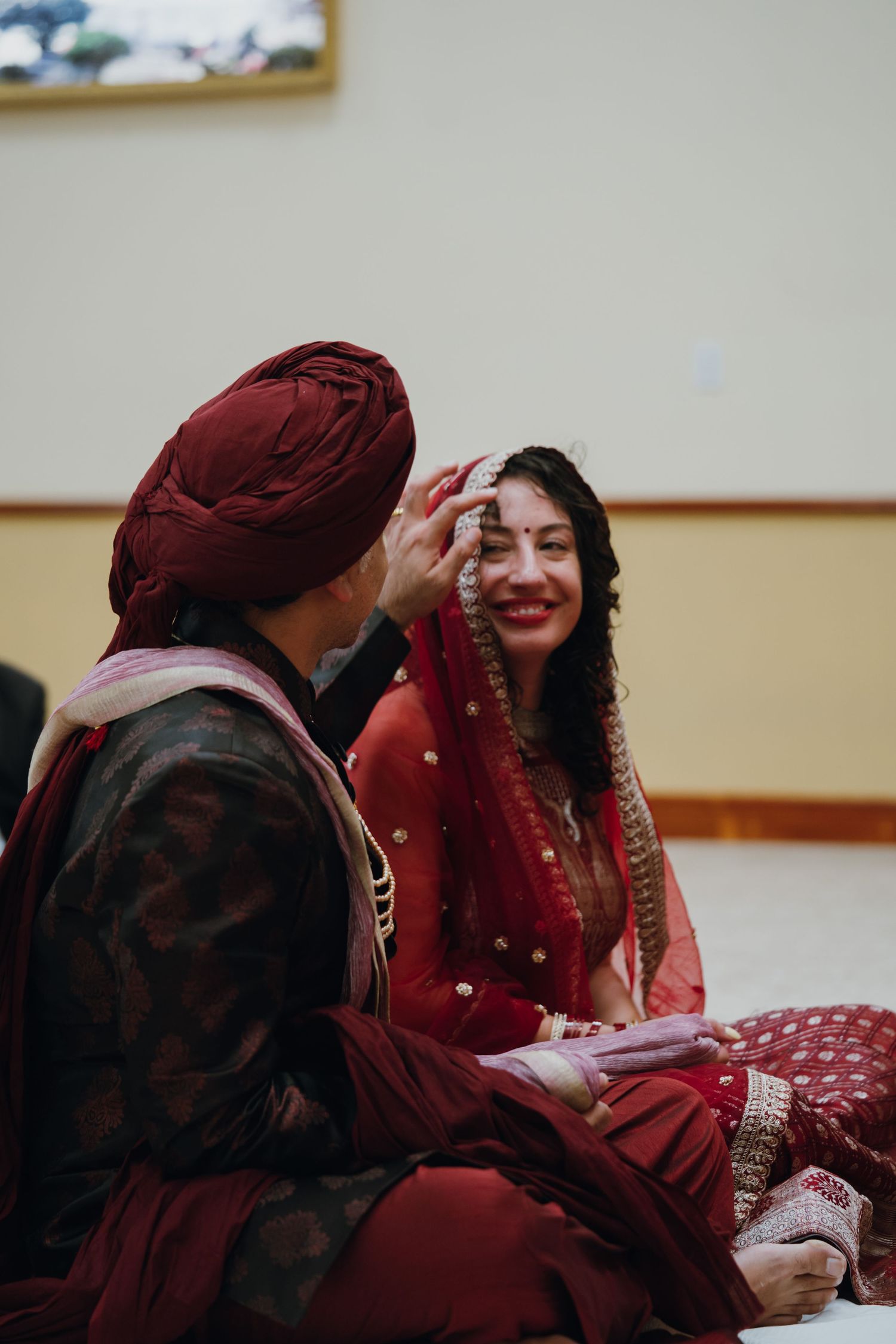 Groom in burgundy turban applies sindoor to smiling bride during traditional Sikh wedding ceremony.