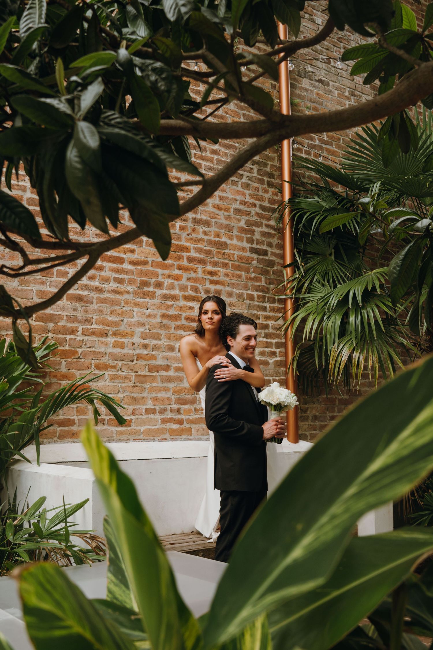 Bride and groom embracing in a lush tropical garden courtyard with exposed brick walls.
