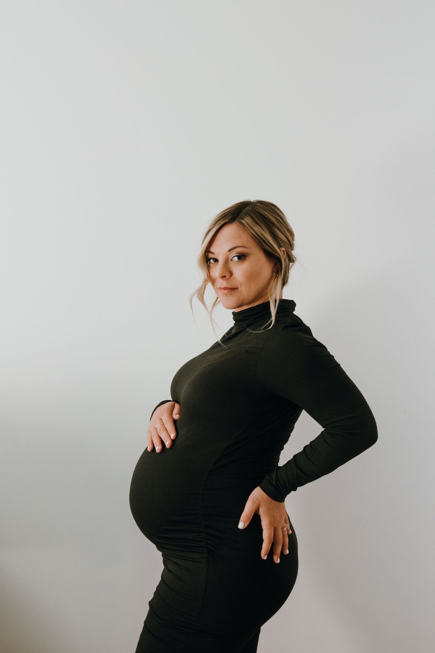 Pregnant woman in black turtleneck dress posing sideways, hands on baby bump.