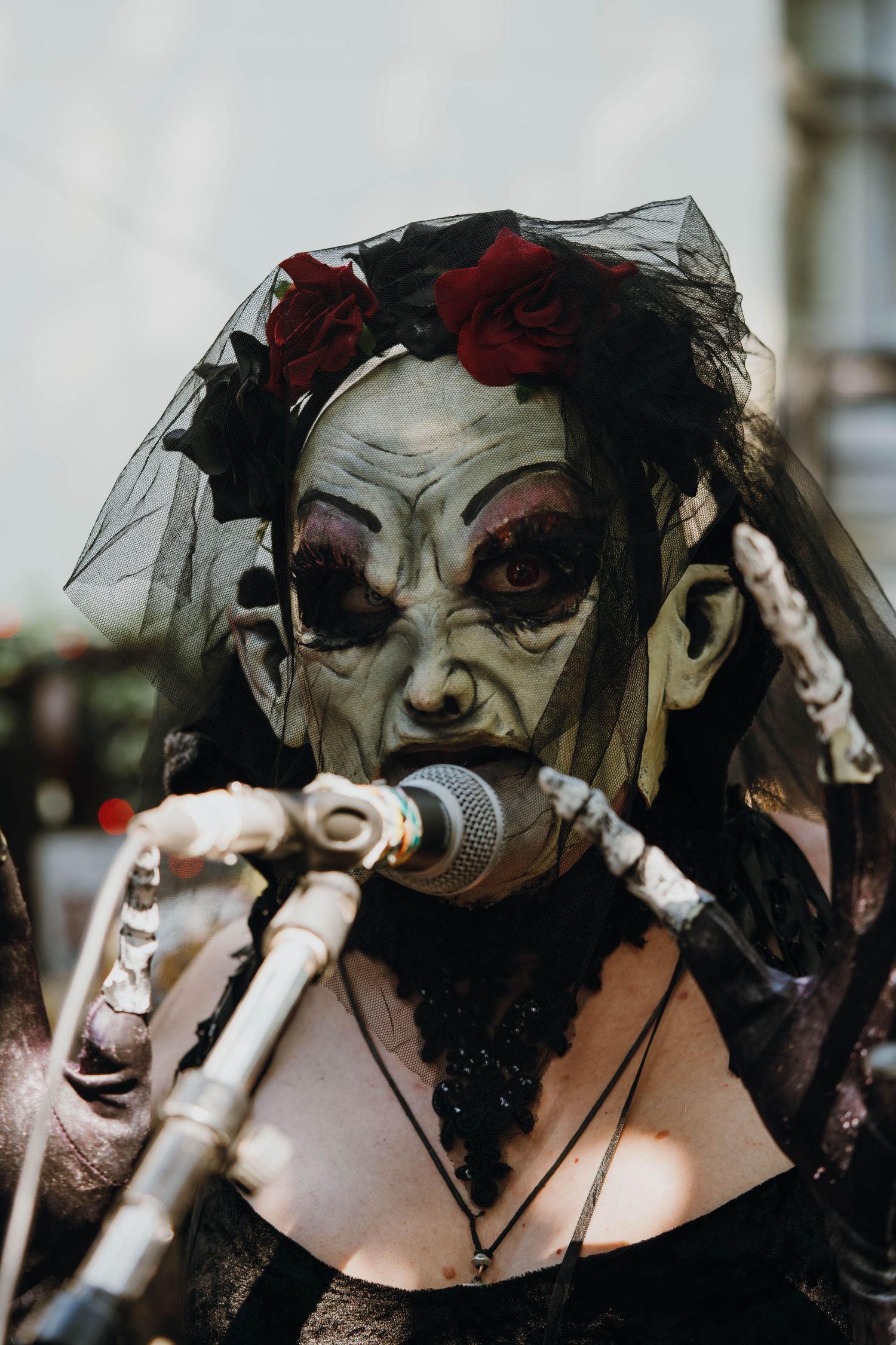Performer wearing a creepy witch mask with red roses and black veil sings into a microphone outdoors.