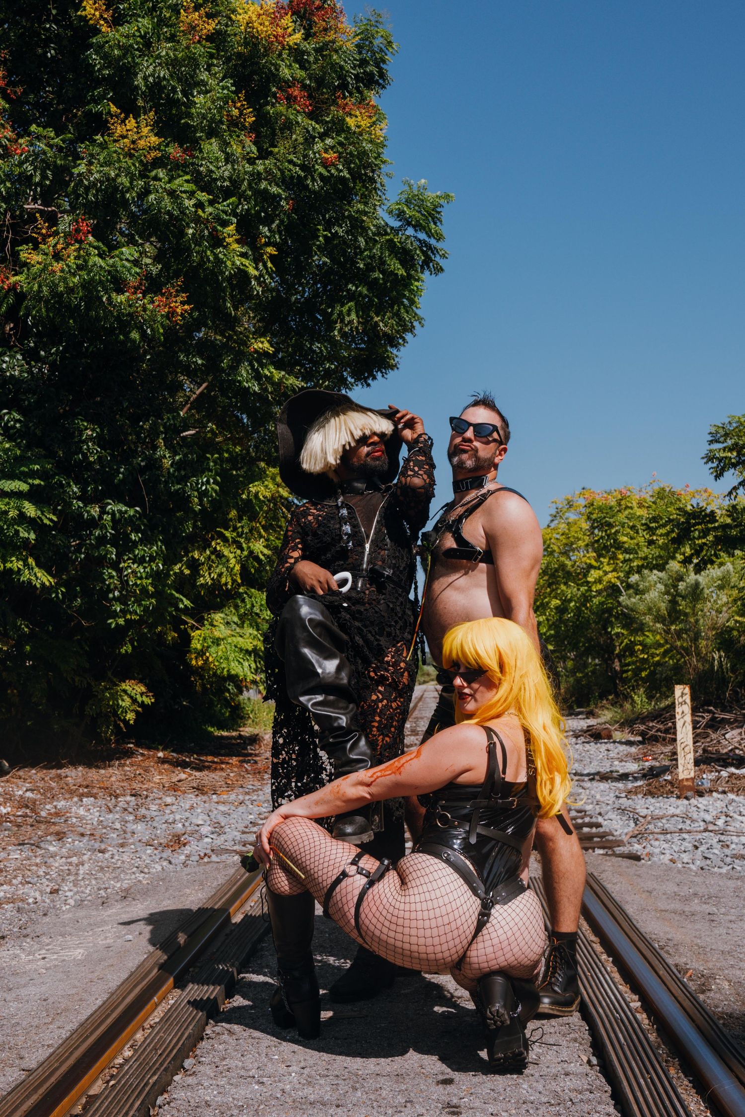 Three people in bold costumes posing on railroad tracks surrounded by lush trees.