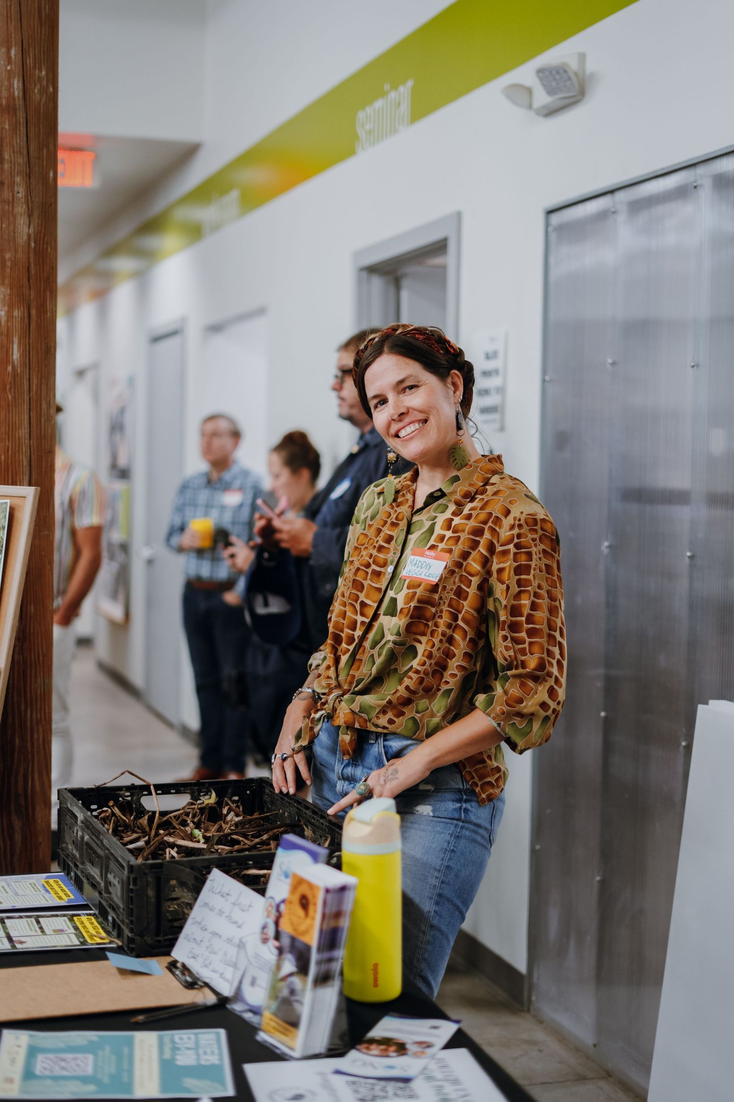 Smiling woman standing at an informational table with a crate of natural materials at an event.