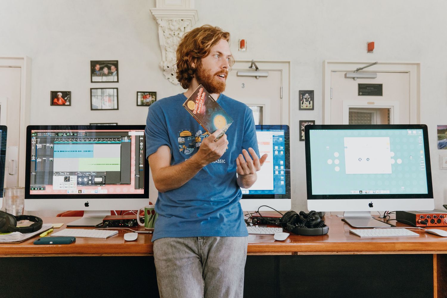 Man in blue t-shirt standing between multiple computer monitors in a creative workspace studio.