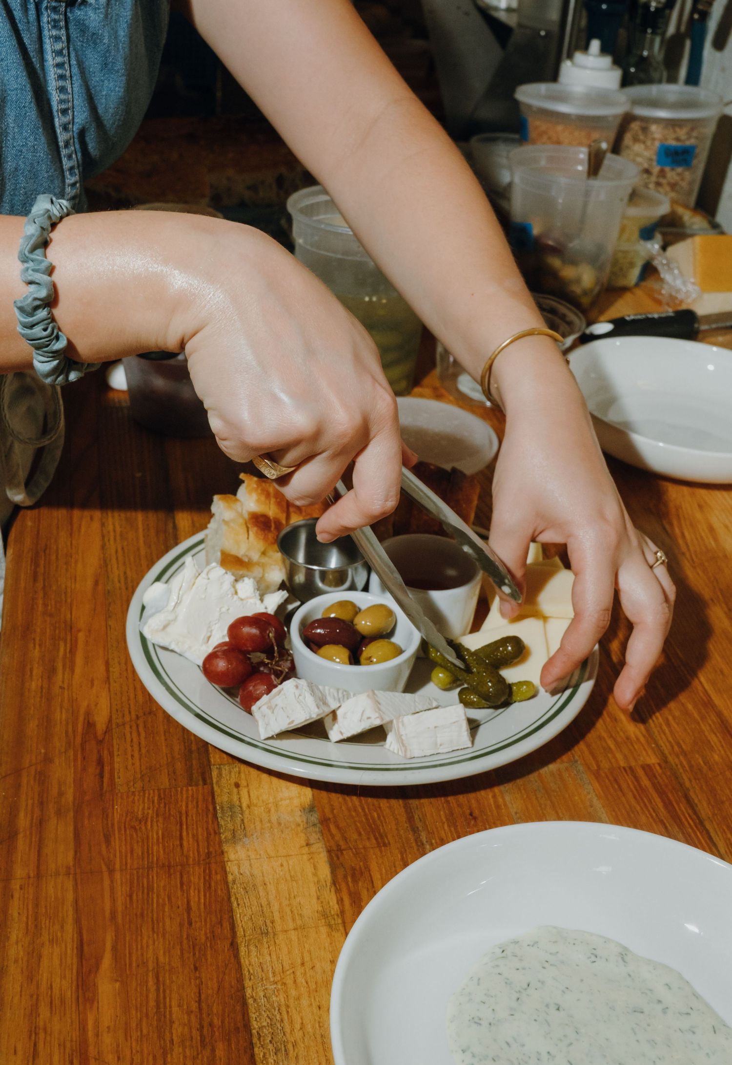 Person serving from a cheese and charcuterie plate with olives, pickles, and bread at a restaurant.