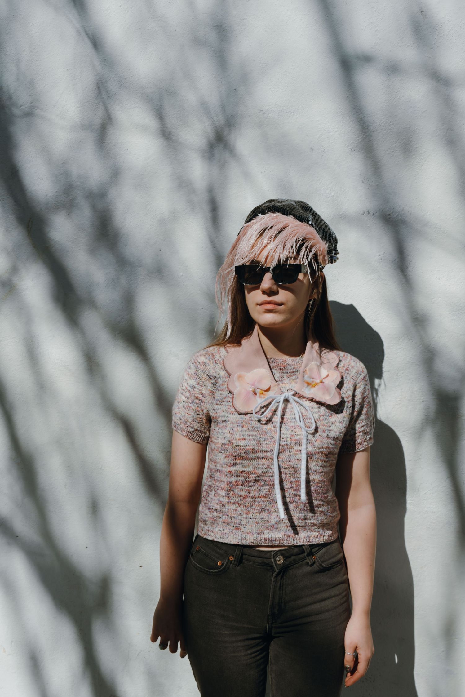 Woman with pink-tipped hair and sunglasses standing against a white wall with tree branch shadows.