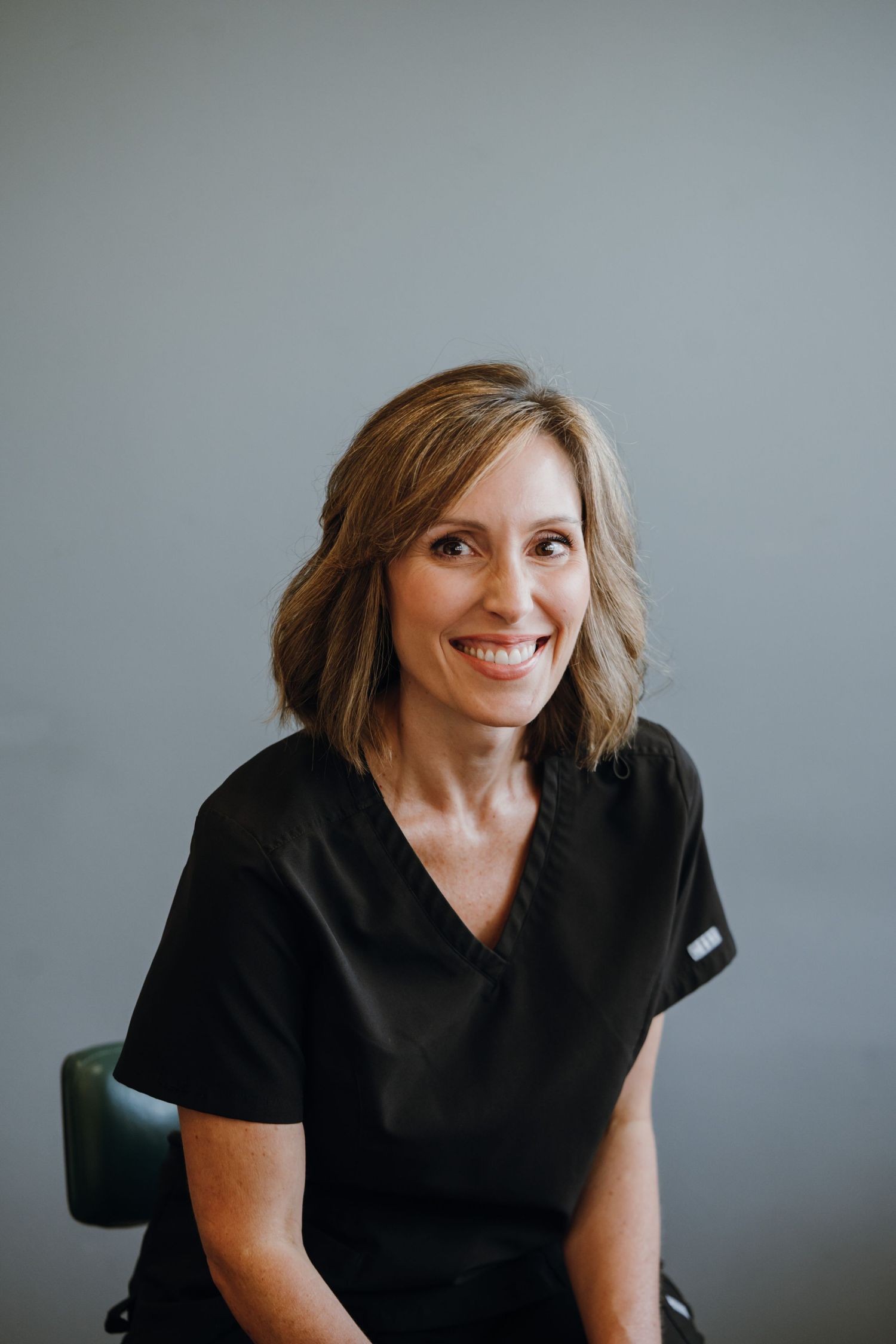 Smiling woman with shoulder-length brown hair wearing black scrubs seated against gray background.