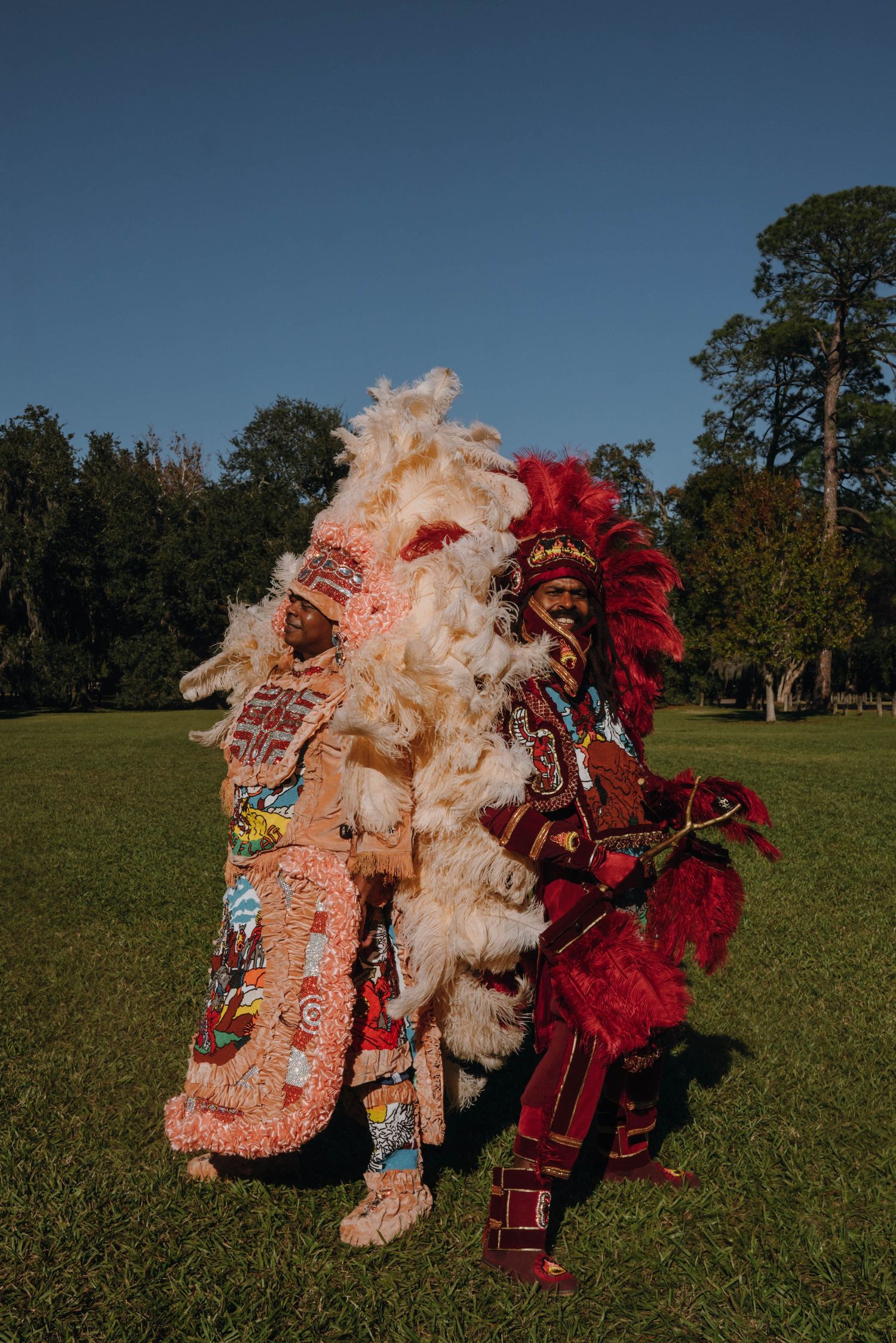Two Mardi Gras Indians in elaborate beaded costumes with feathered headdresses stand together on green grass.