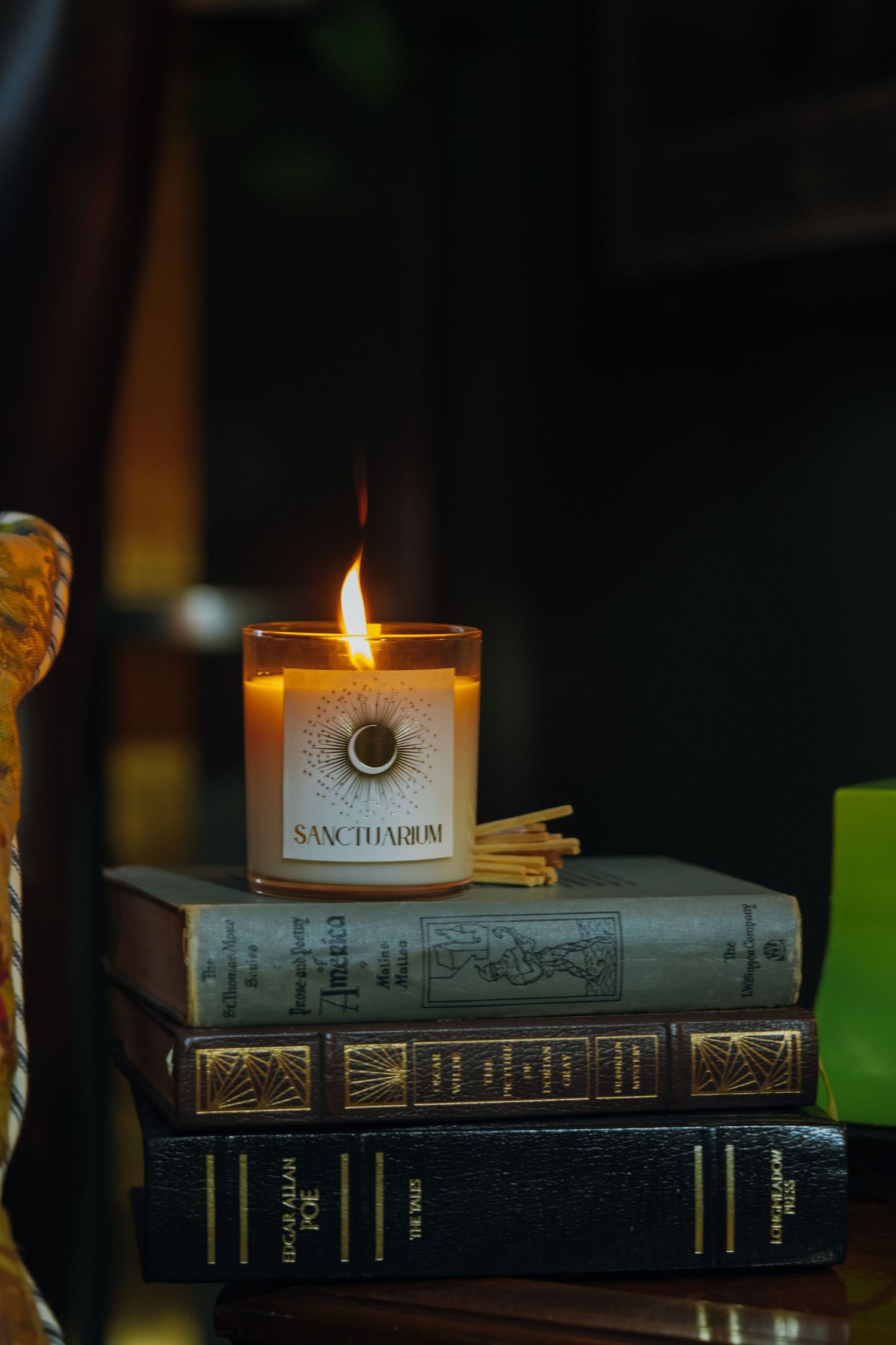 Lit Sanctuarium candle on stack of vintage books with matches in moody dark setting.