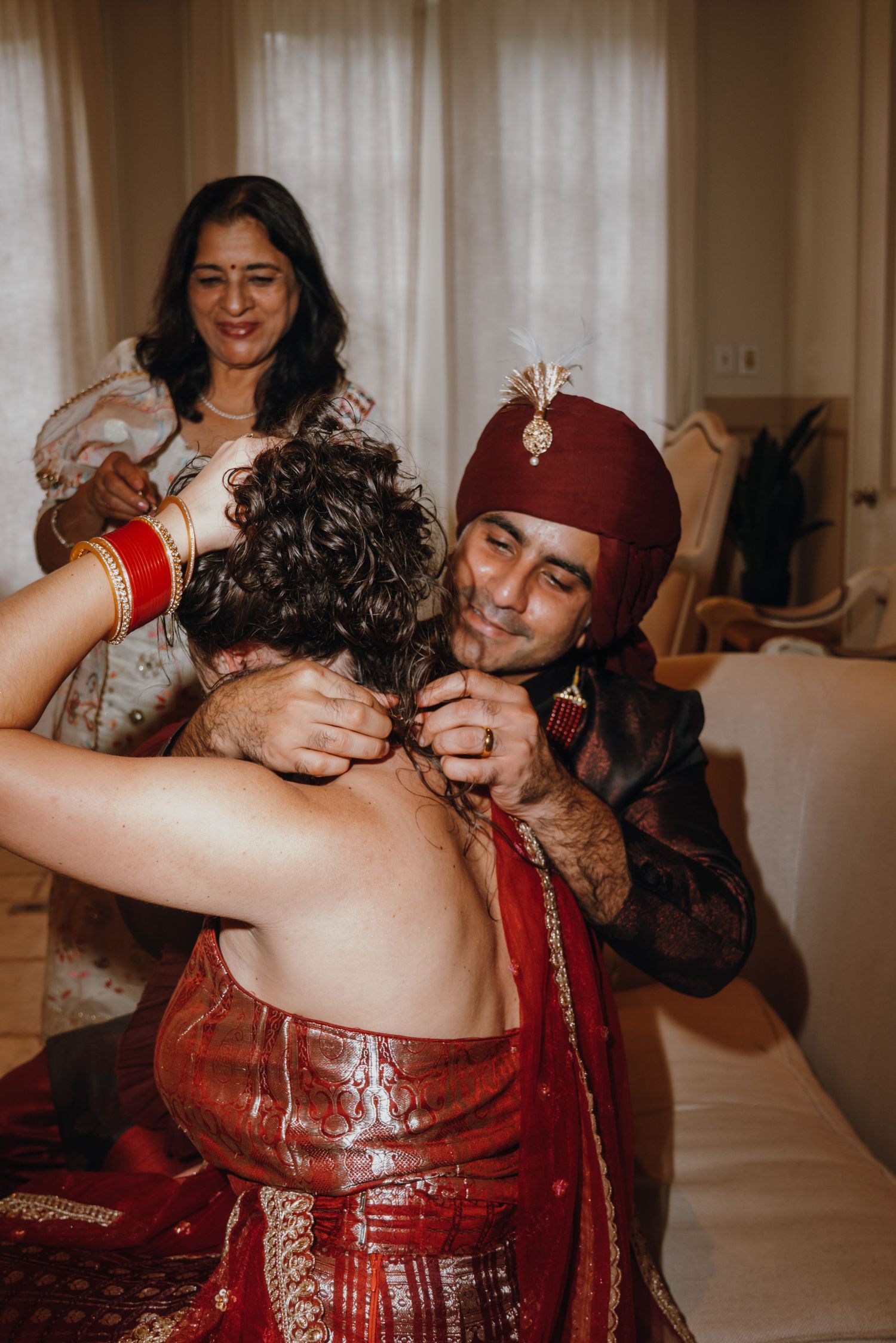 Groom in red turban sharing a tender moment with bride while woman assists with outfit.