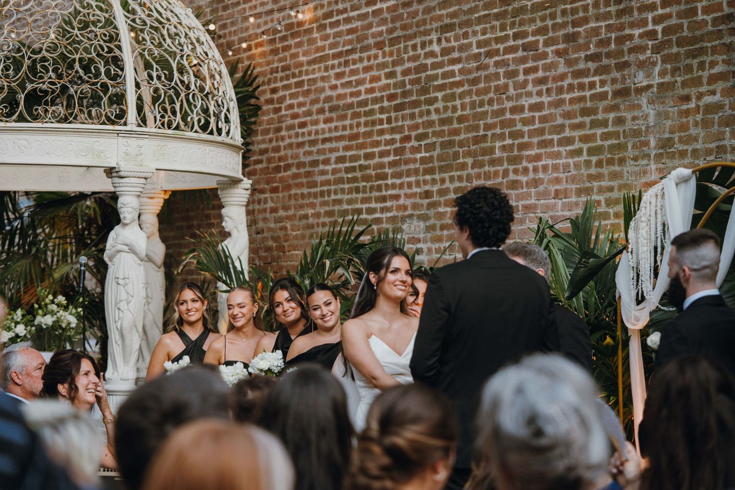 Bride smiles at groom during outdoor wedding ceremony near a white gazebo and brick wall.