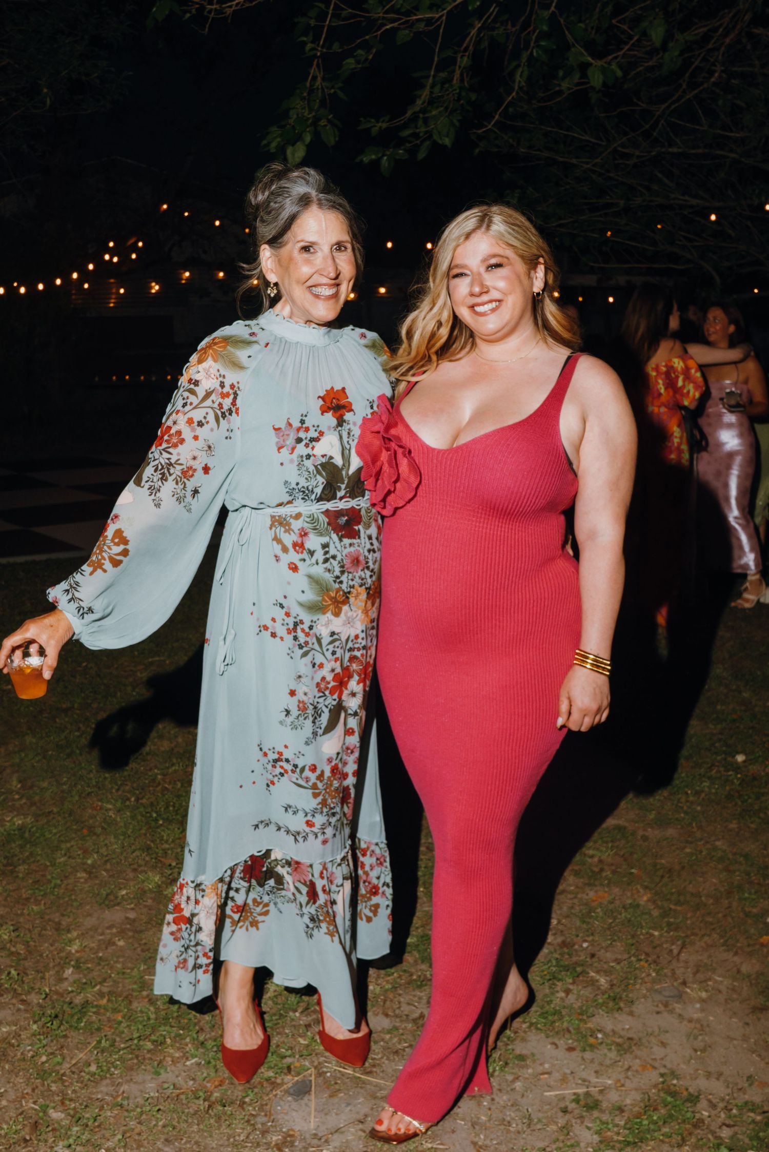 Two women pose together at outdoor wedding reception, one in floral dress and one in red gown.
