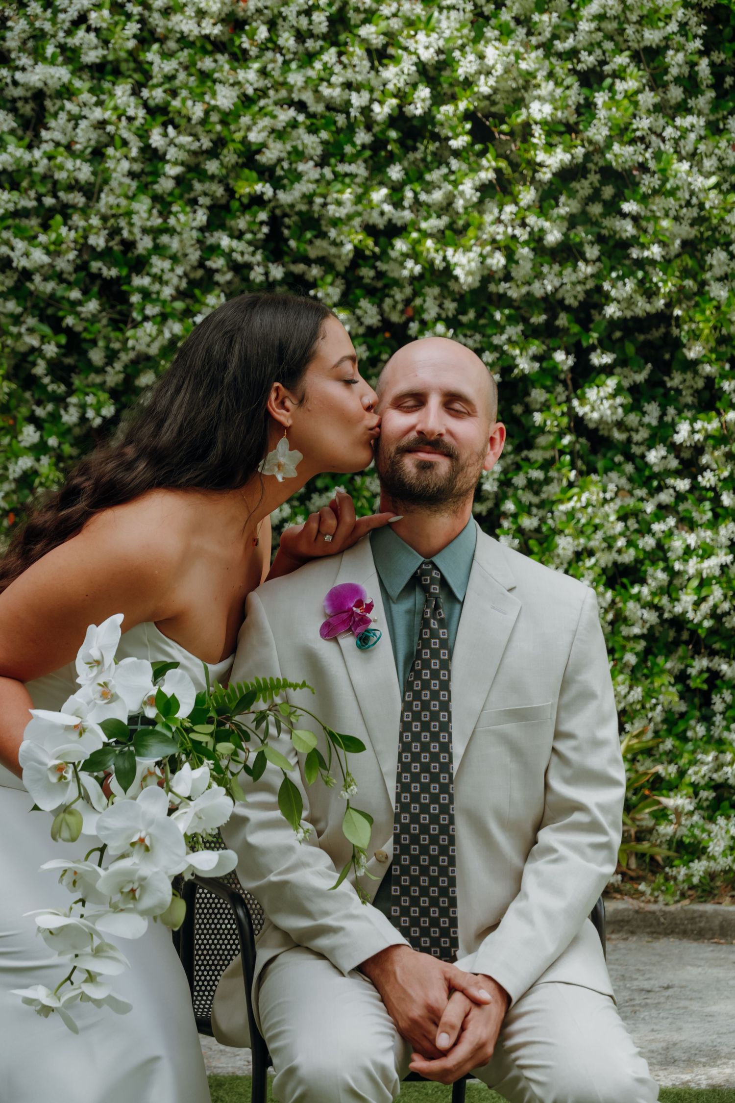 Bride kisses groom on cheek as he sits before lush white floral wall holding her bouquet.