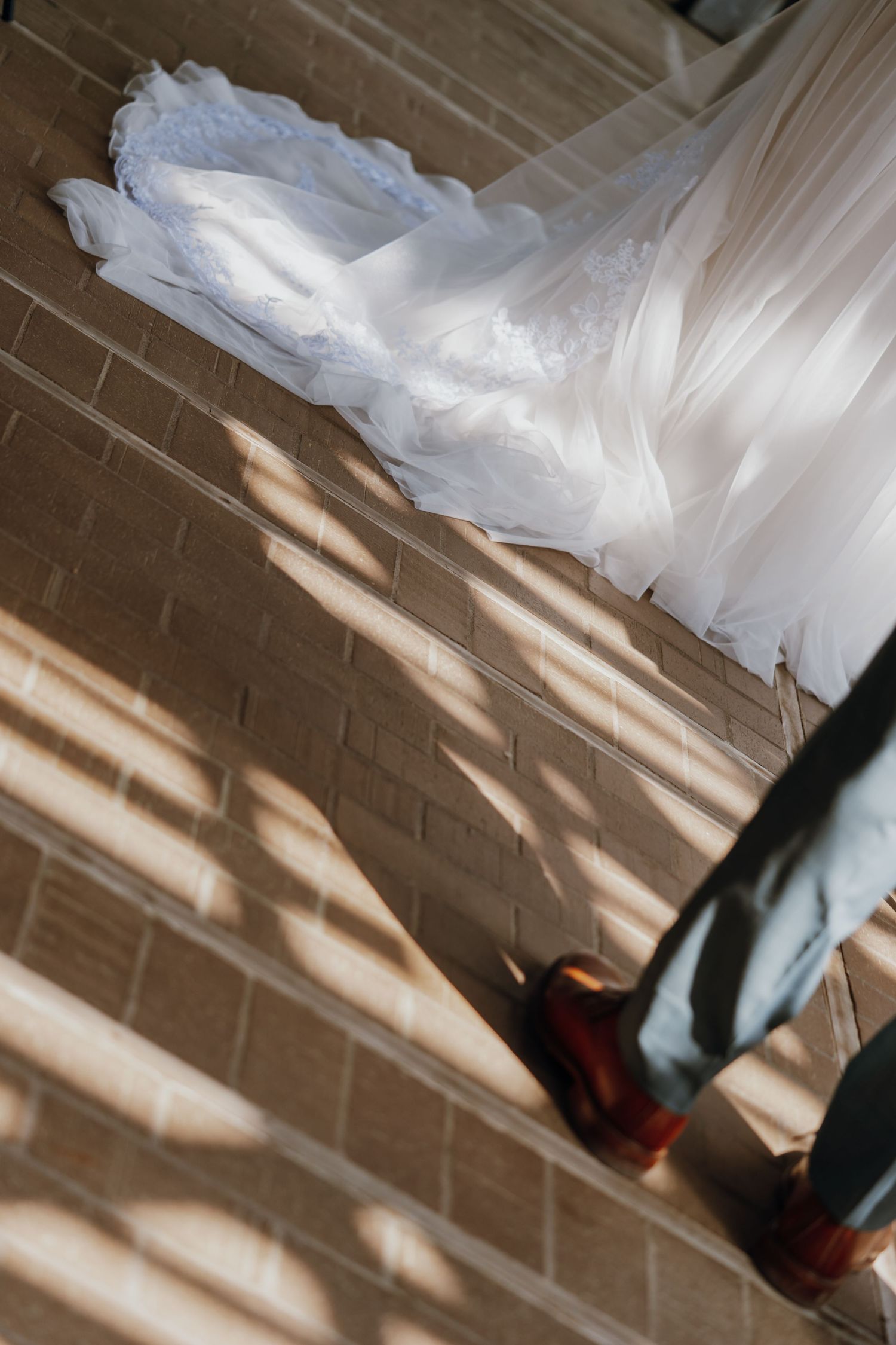 Close-up of elegant white wedding dress train on brick floor with groom's brown leather shoes beside it.