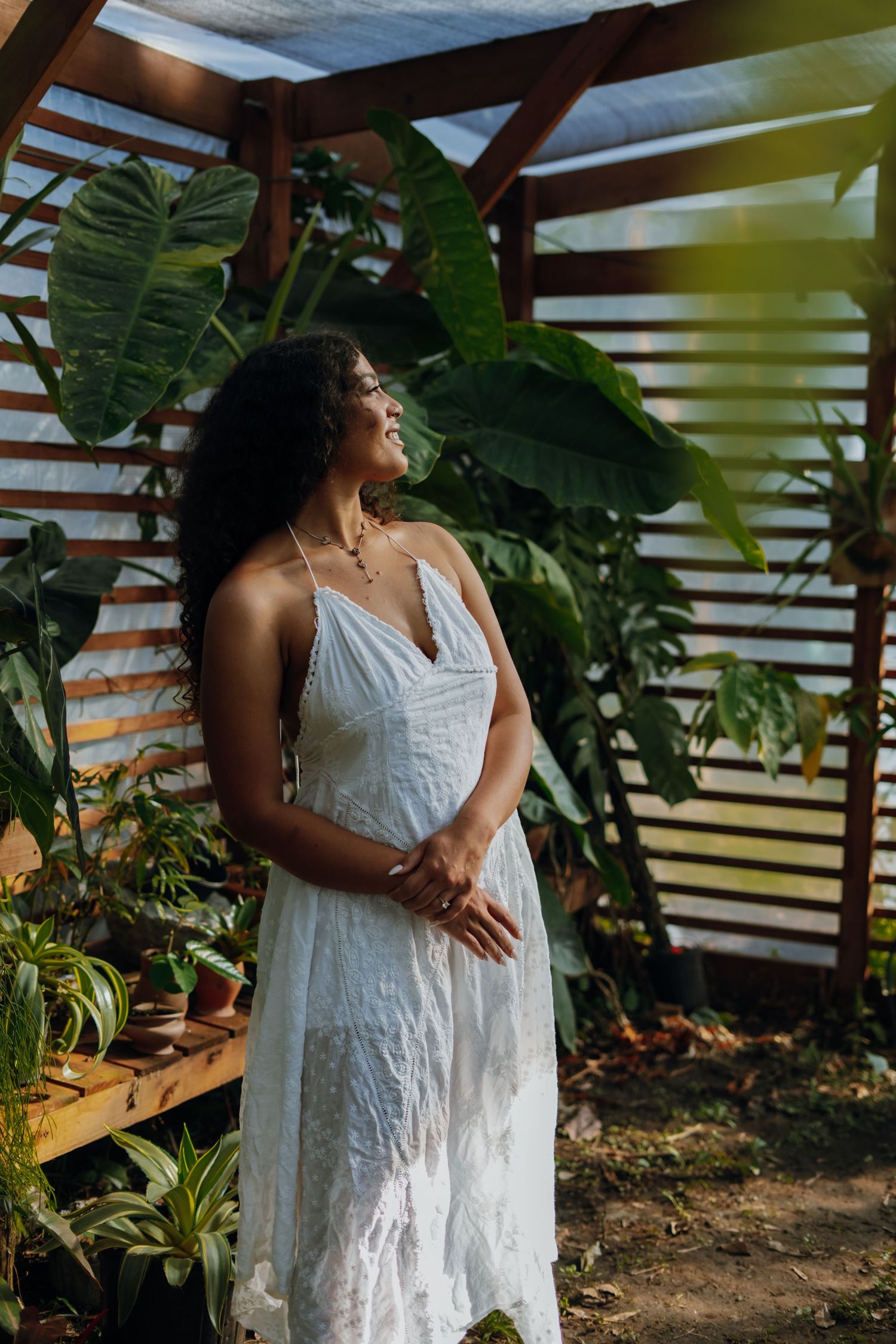 A curly-haired woman in a white lace dress gazes upward while standing among tropical plants in a greenhouse.