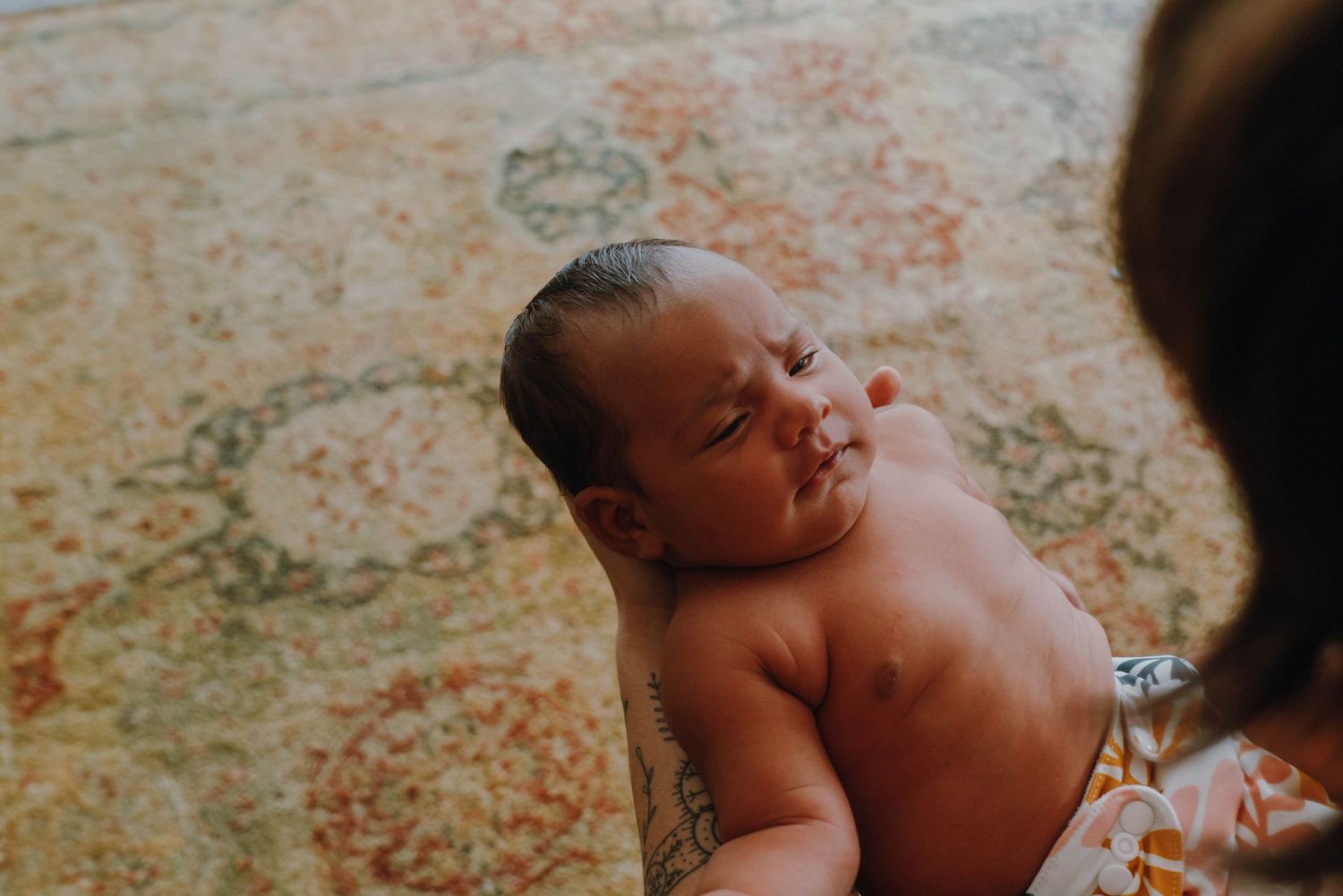 Newborn baby smiling while being held by tattooed parent over colorful patterned carpet. New Orleans, Louisiana, Mississippi