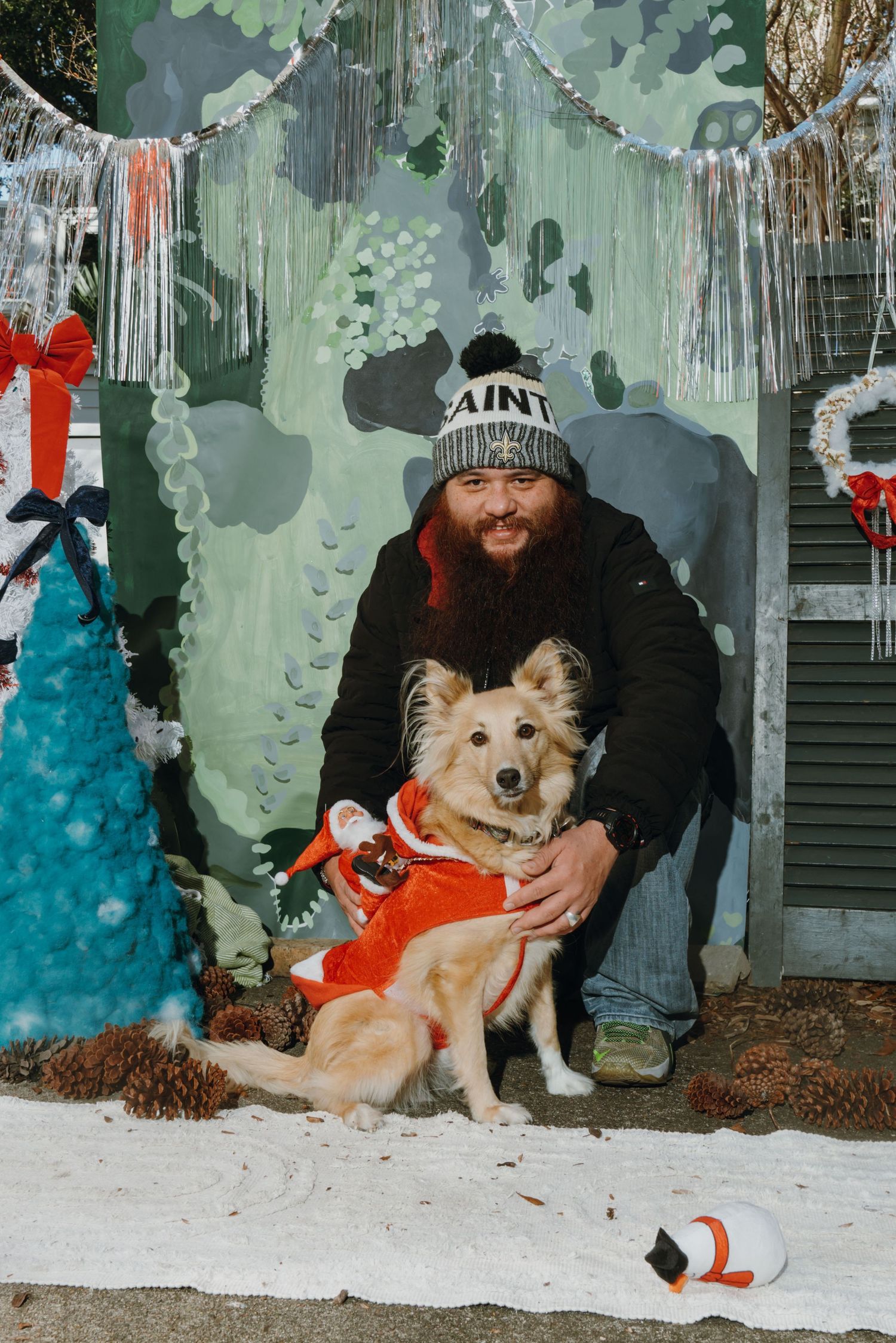 Bearded man in Saints beanie poses with a dog in Santa outfit amid Christmas decorations and artificial snow.