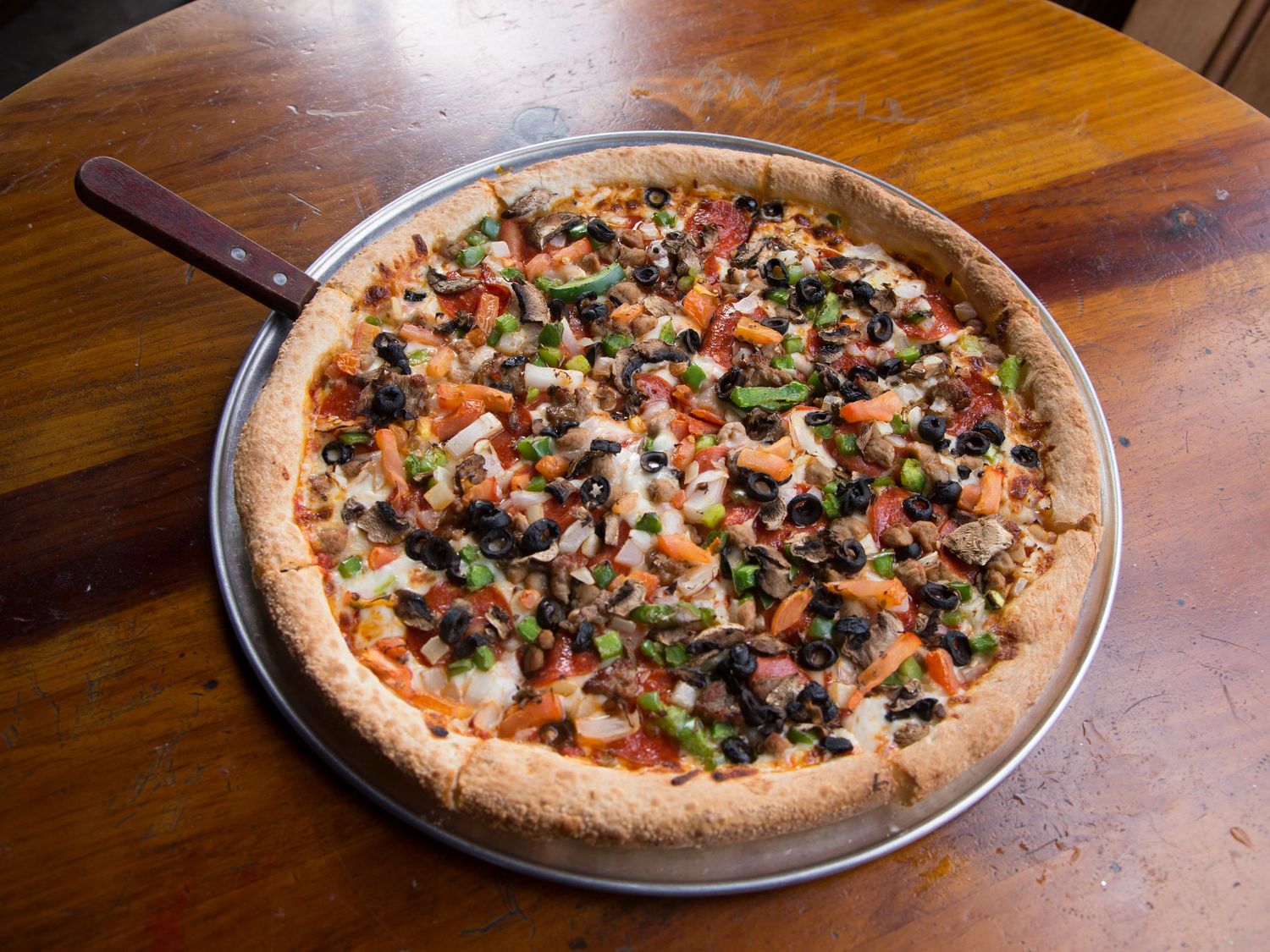 A loaded supreme pizza with black olives, peppers, onions, and mushrooms on a silver pan atop a wooden table.