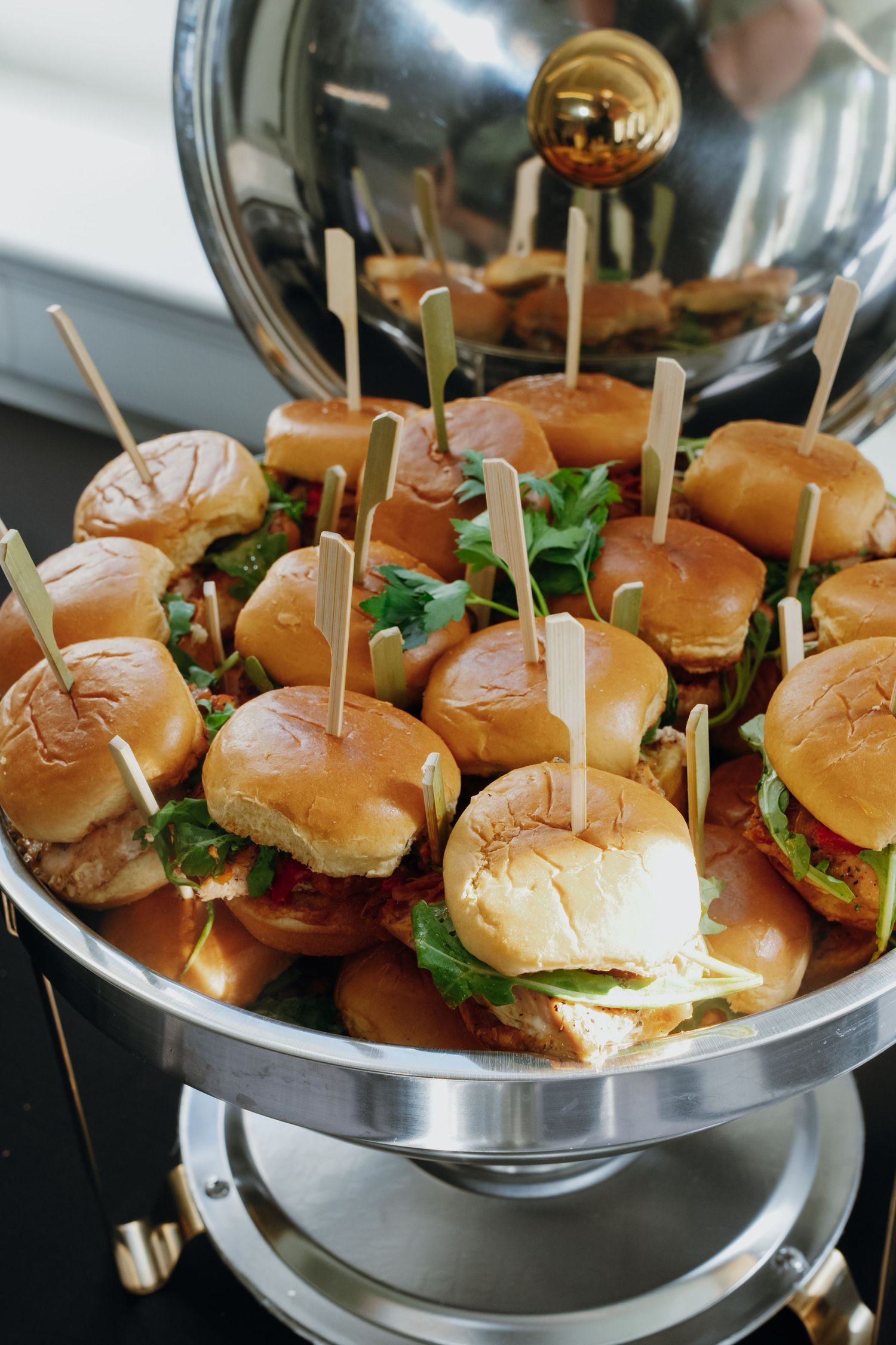 Tray of mini sliders with toothpick garnishes served in a silver chafing dish at catered event.