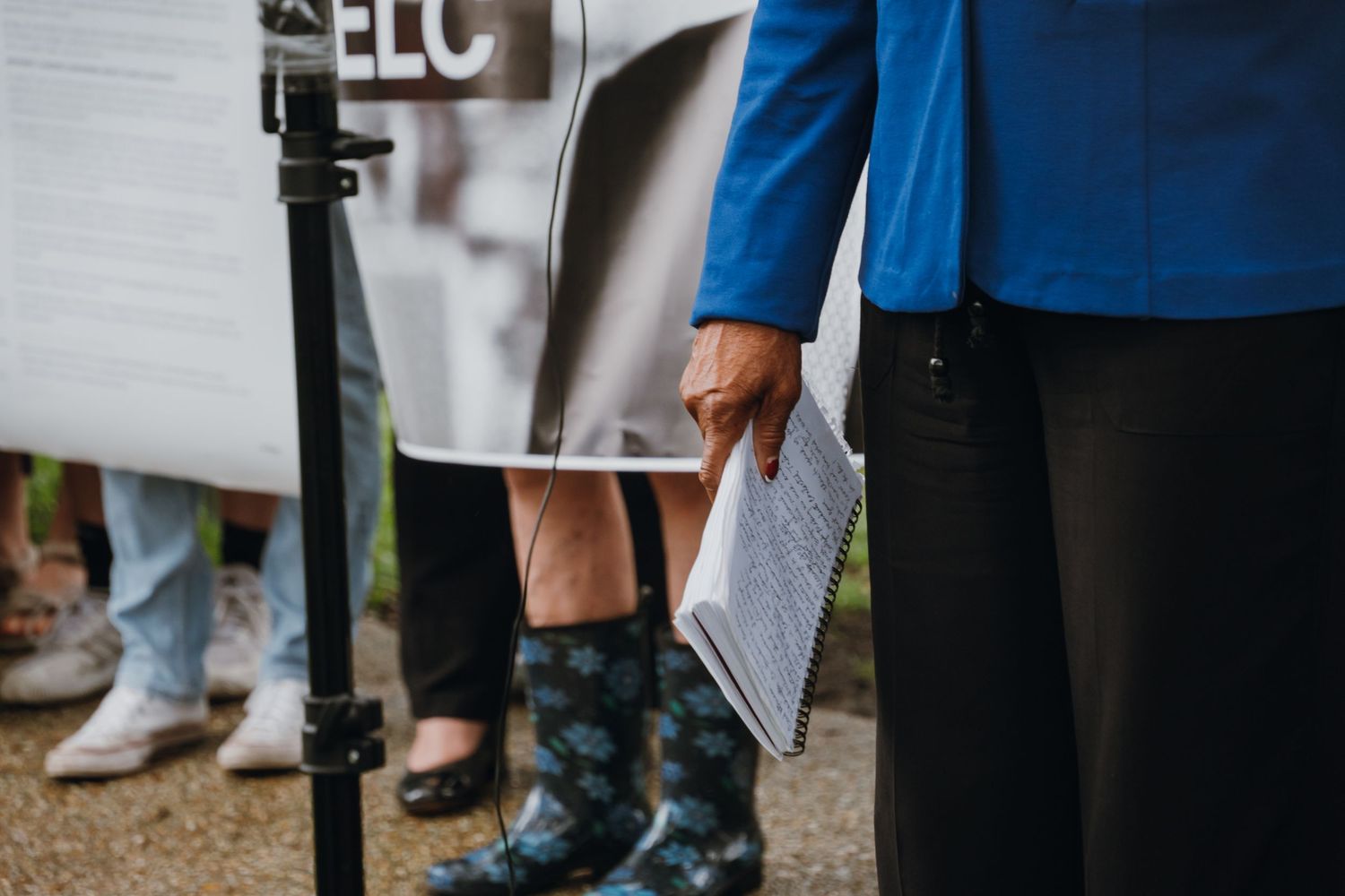 People standing outdoors holding papers and signs, wearing rain boots and blue jacket at a public gathering.